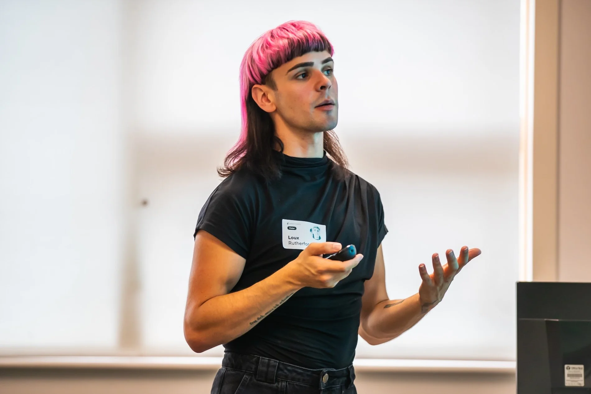 A person with pink and black hair giving a presentation in front of a whiteboard, holding a remote and wearing a black shirt with a name tag that reads 'Loux Rutherford'.