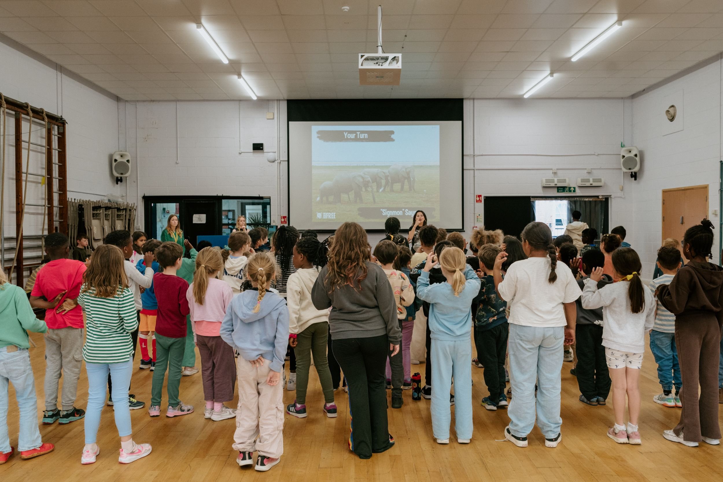 A large group of children gathered in a school gymnasium watching a presentation projected on a screen. The children are standing and facing the screen, which displays the words "Your Turn" above an image of elephants. The room has white walls, a wooden floor, and a ceiling with fluorescent lights.