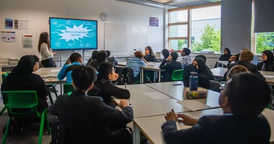 Classroom filled with students and a teacher at the front, presenting a slide on a screen about campaign visuals, with large windows showing trees outside.