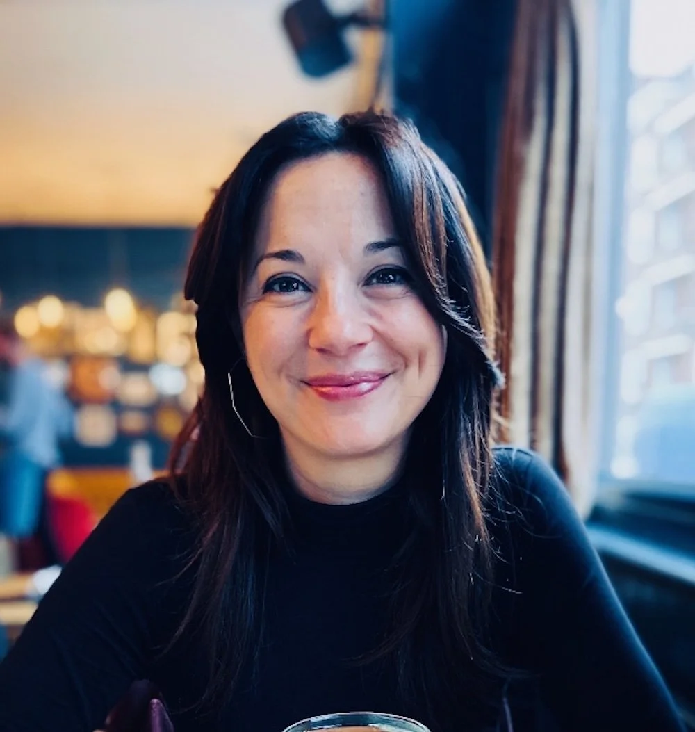 A smiling woman with dark hair sitting in a restaurant with a window to her right and warm lighting in the background.