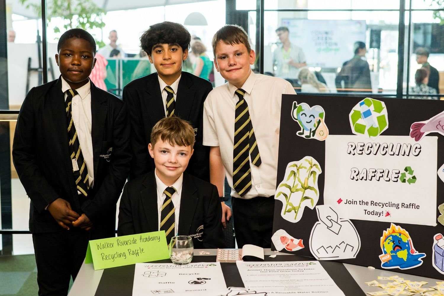 Four young boys in school uniforms standing behind a table at a recycling raffle event, with a poster titled 'Recycling Raffle' and various illustrations related to recycling.