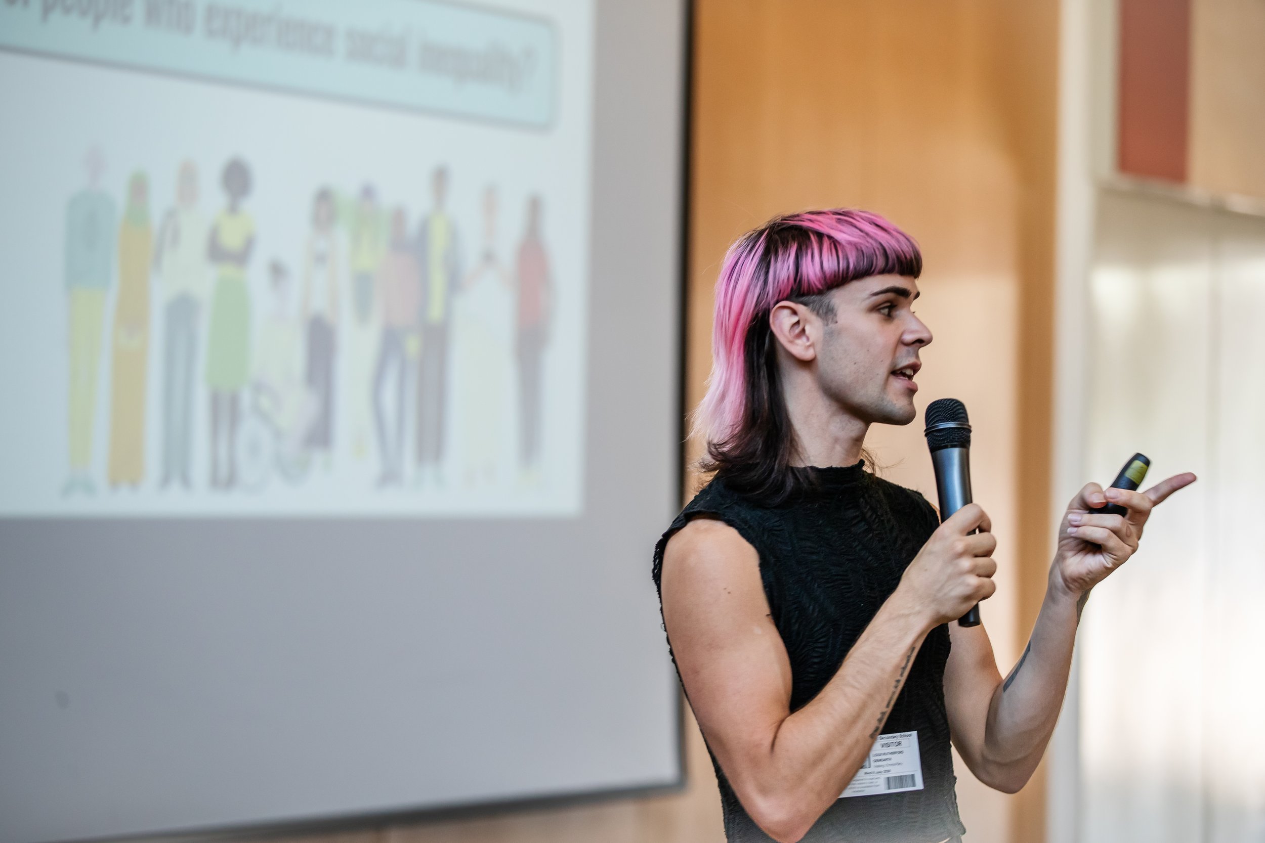 A person with pink and black hair giving a presentation in front of a whiteboard, holding a remote and wearing a black shirt with a name tag that reads 'Loux Rutherford'.