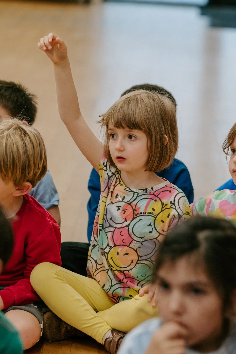 A young girl with short brown hair wearing a colorful shirt and yellow pants sits with legs crossed, raising her hand in a classroom or group setting, surrounded by other children.