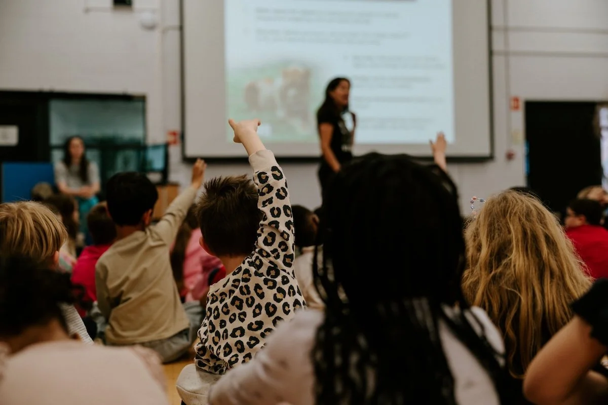 Children sitting on the floor of a classroom, with some raising their hands, facing a woman standing at the front in front of a projected presentation.