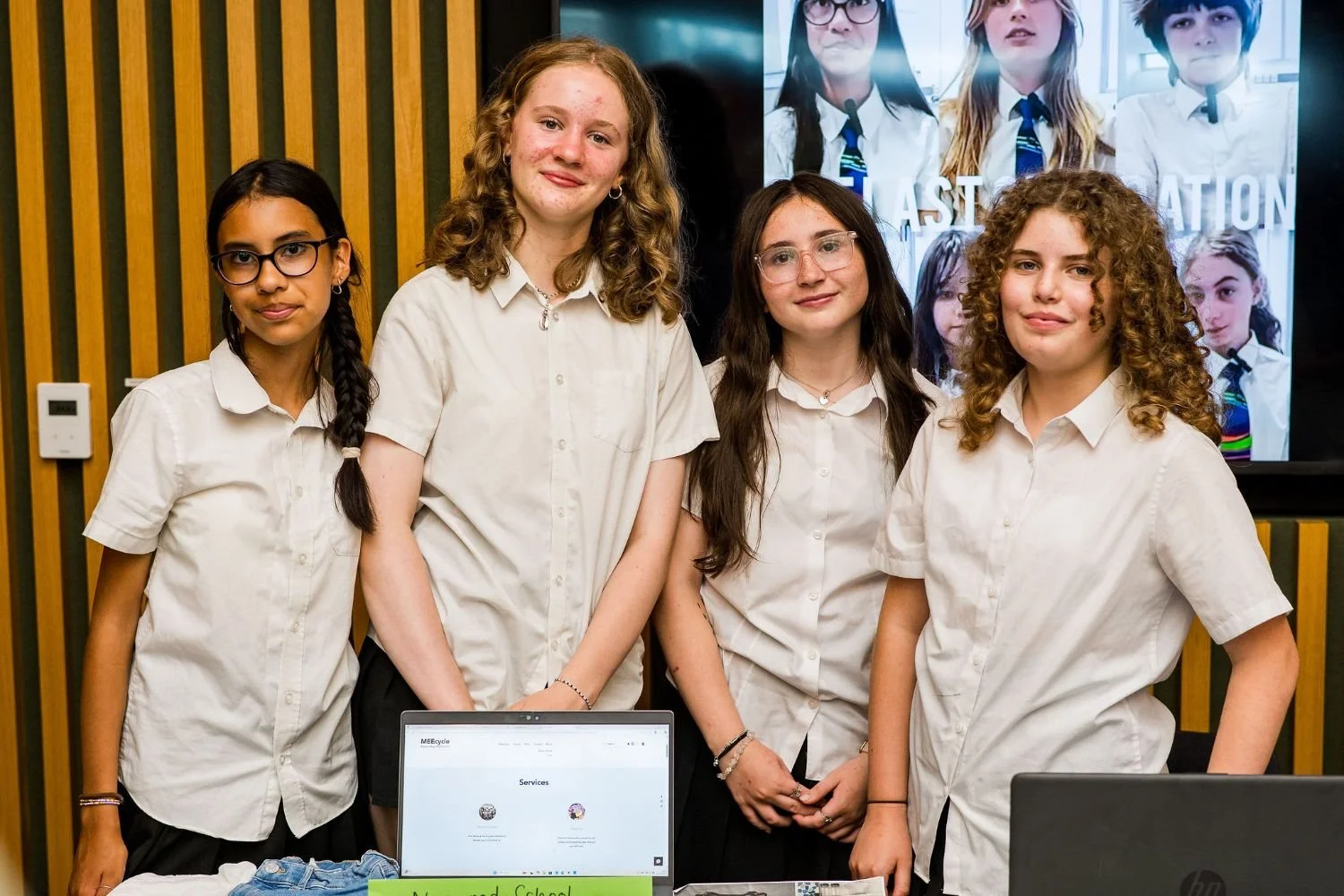 Four girls in white shirts standing behind a table with a laptop, at a school presentation or event, in front of a large screen displaying images of other students.