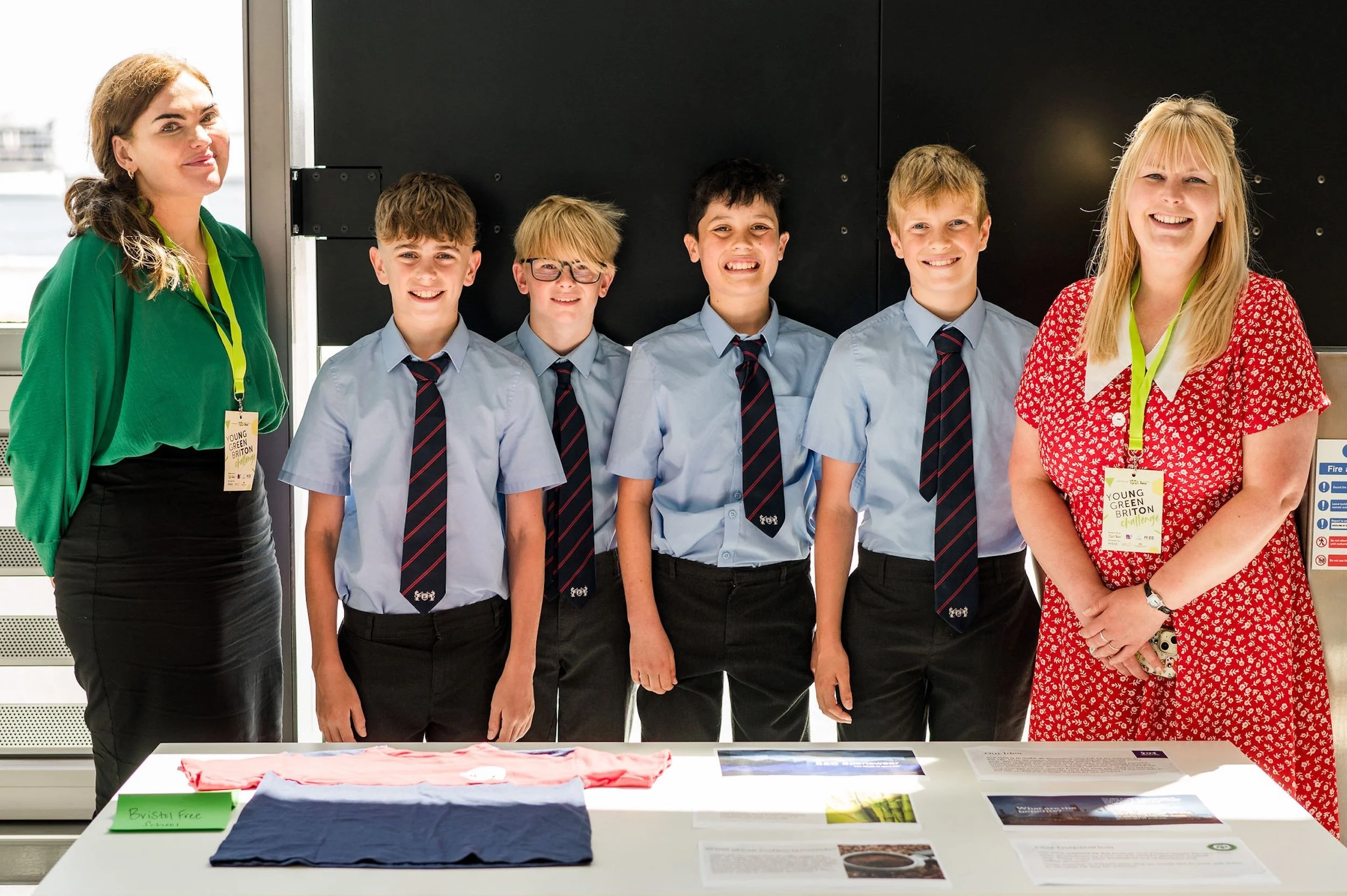 Group of five children and two teachers standing behind a table displaying folded clothes, with a black background.