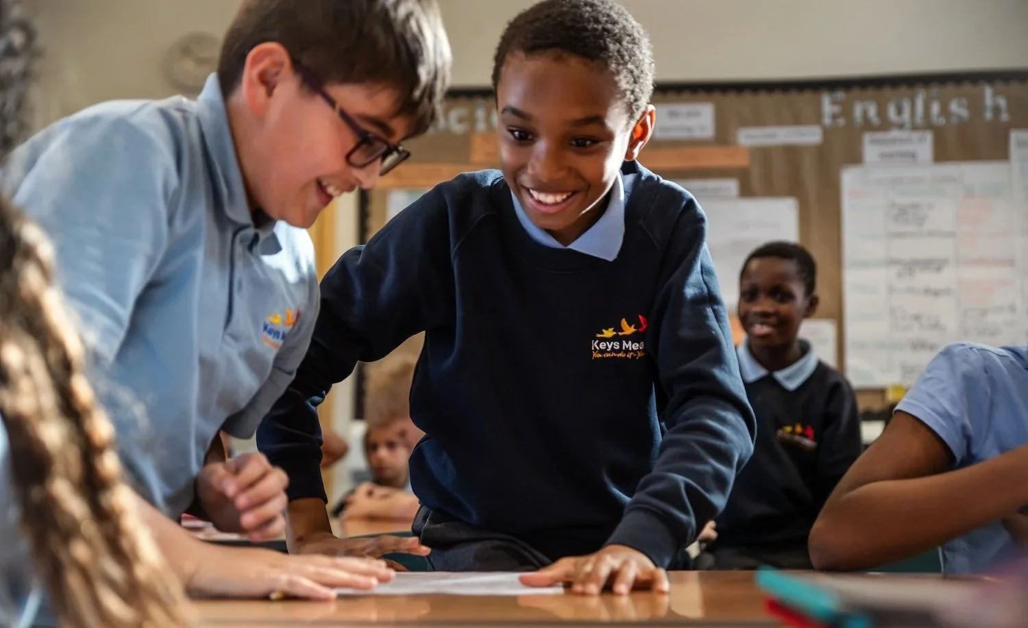 Two smiling boys interacting with a teacher or adult in a classroom, with other students in the background working at desks.