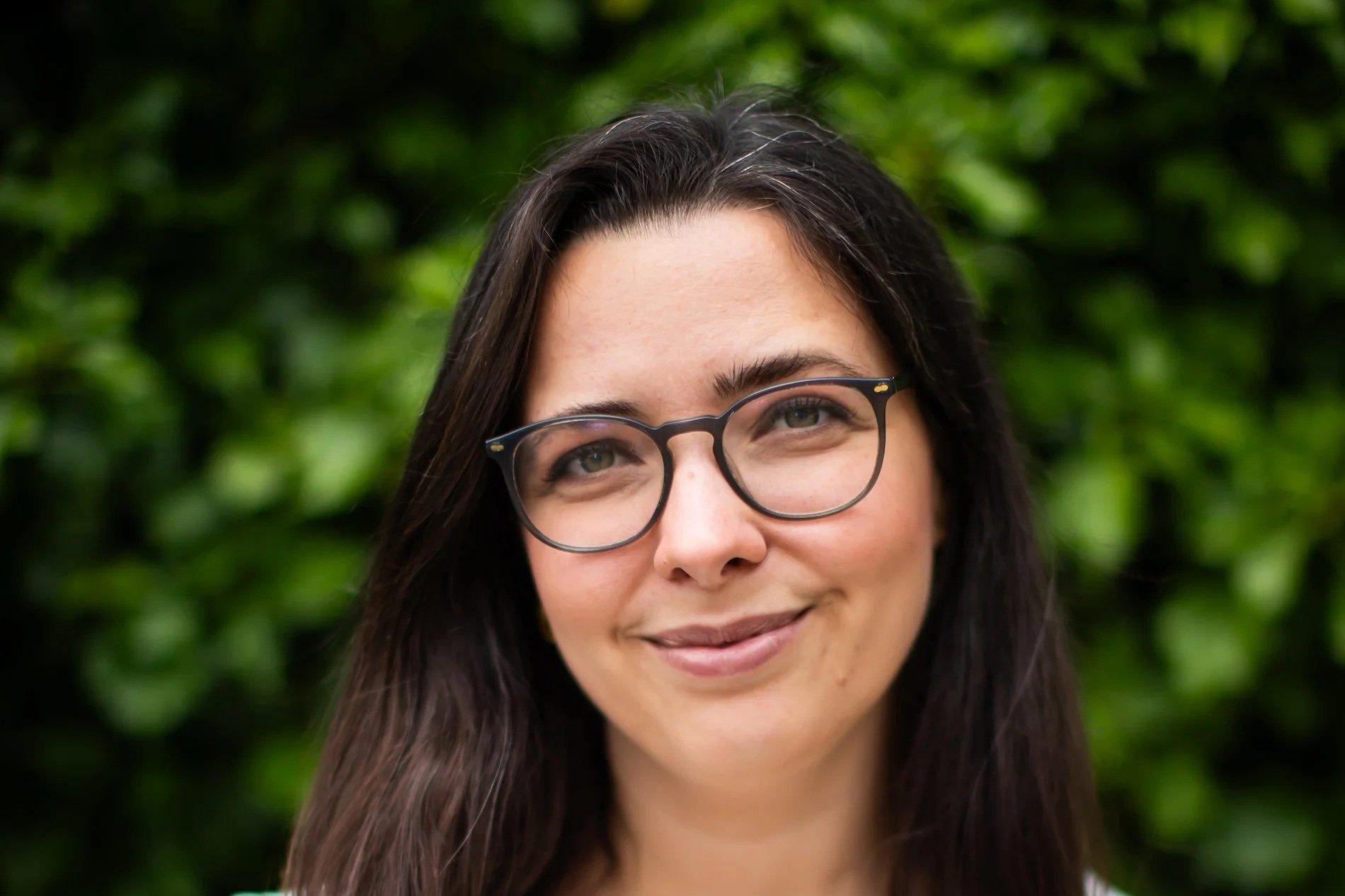 Close-up of a smiling woman with long dark hair, wearing glasses, against a blurred green leafy background.