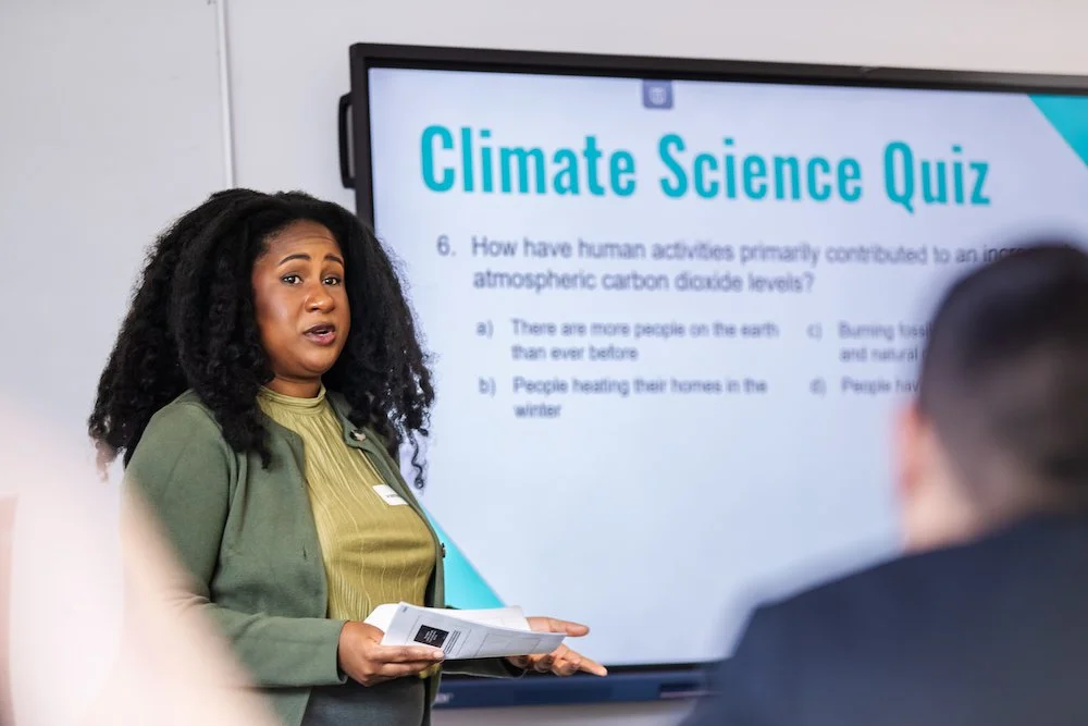 A woman presenting a climate science quiz on a large screen in a classroom or conference room, with students listening.