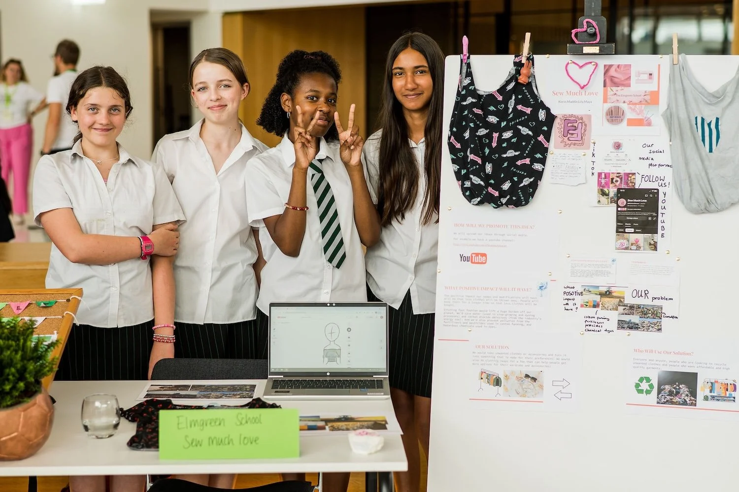 Four girls in school uniforms standing behind a table with a laptop and a poster board, smiling at the camera during a school project presentation.