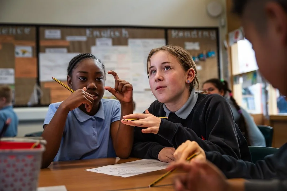 Students in a classroom engaged in a discussion, with two girls in focus and other students in the background.