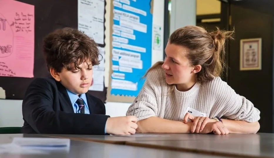 A young boy in a school uniform and a woman sit at a table in a classroom, the boy looks upset and the woman appears to be talking to him to comfort or address him.