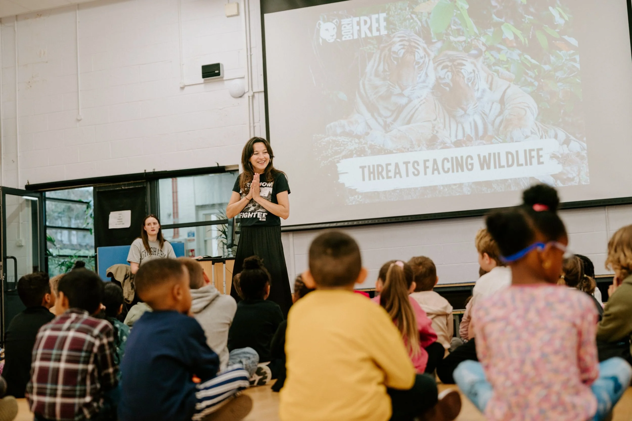 Young woman with long dark hair, smiling, standing in front of a classroom full of students, giving a presentation on wildlife threats, with a slide showing two tiger cubs and the text 'Threats Facing Wildlife' projected behind her.