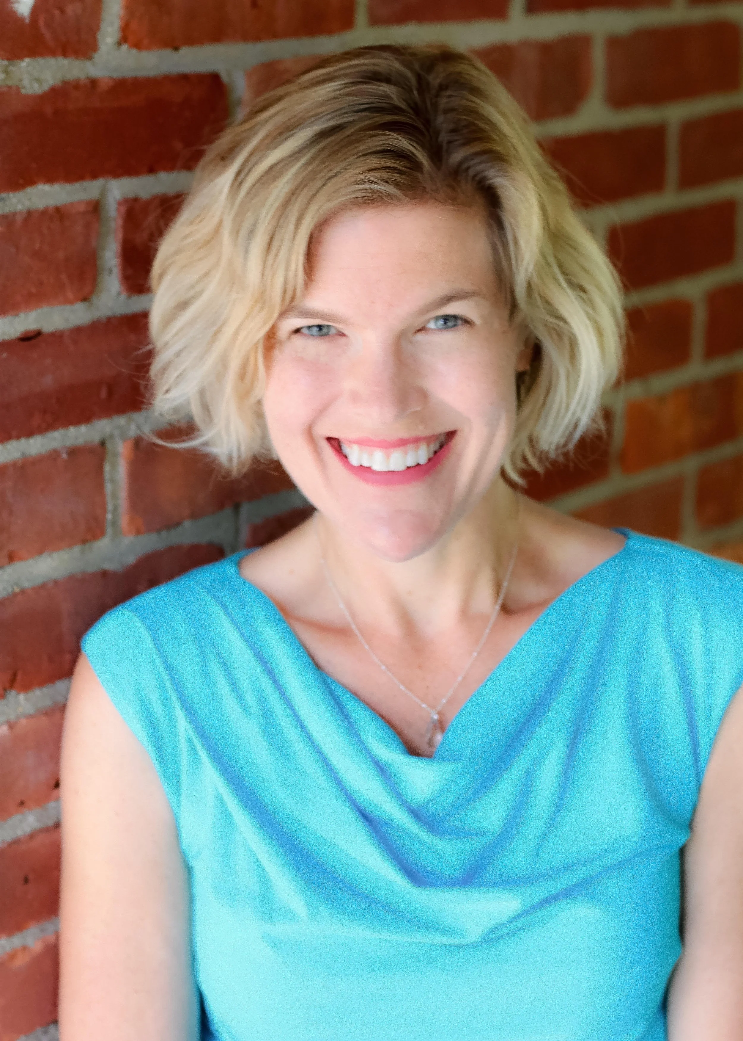 A woman with short blonde hair and blue eyes smiling against a red brick wall, wearing a turquoise dress and a silver necklace.