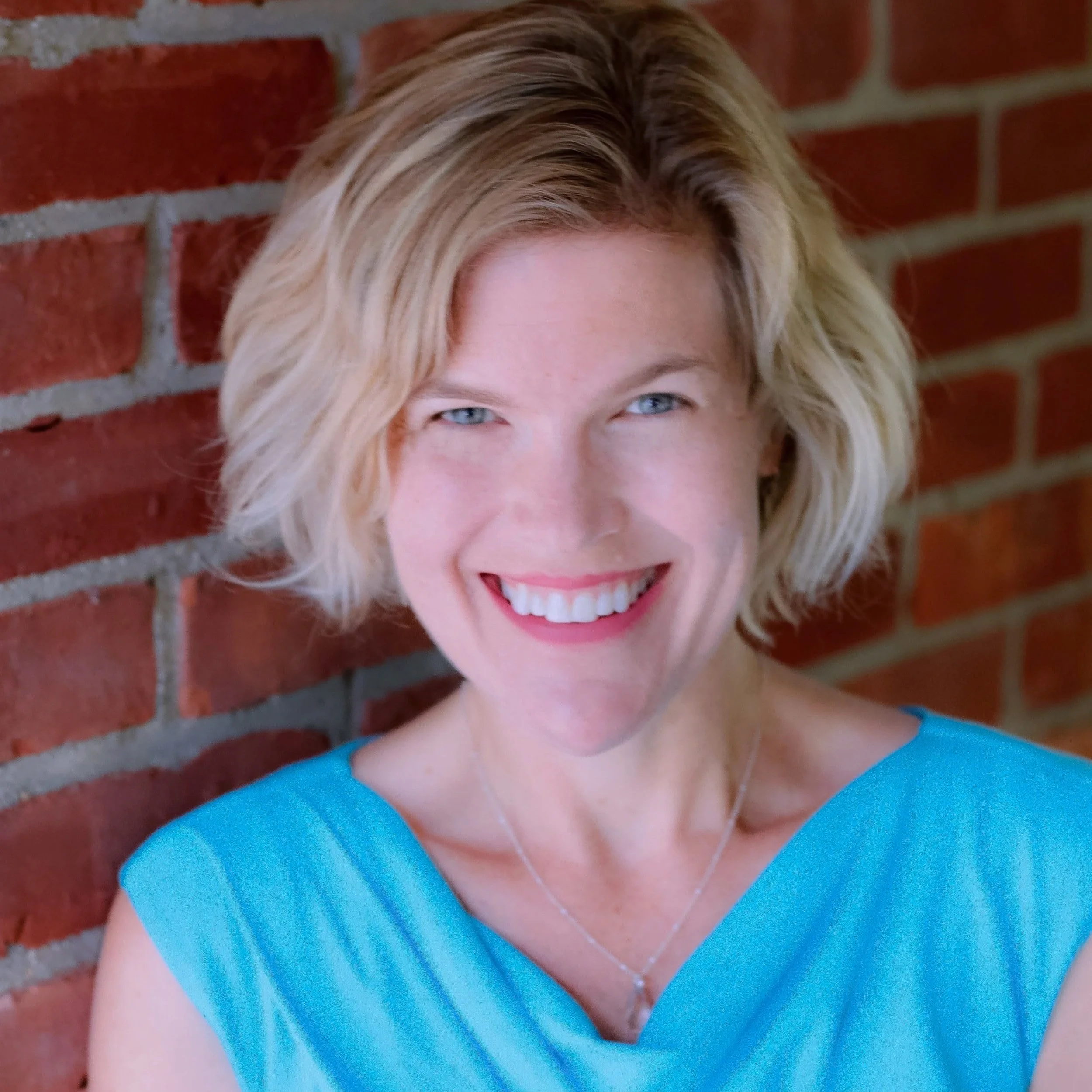 Close-up of a smiling woman with blonde hair and blue eyes, wearing a blue top, standing against a red brick wall.