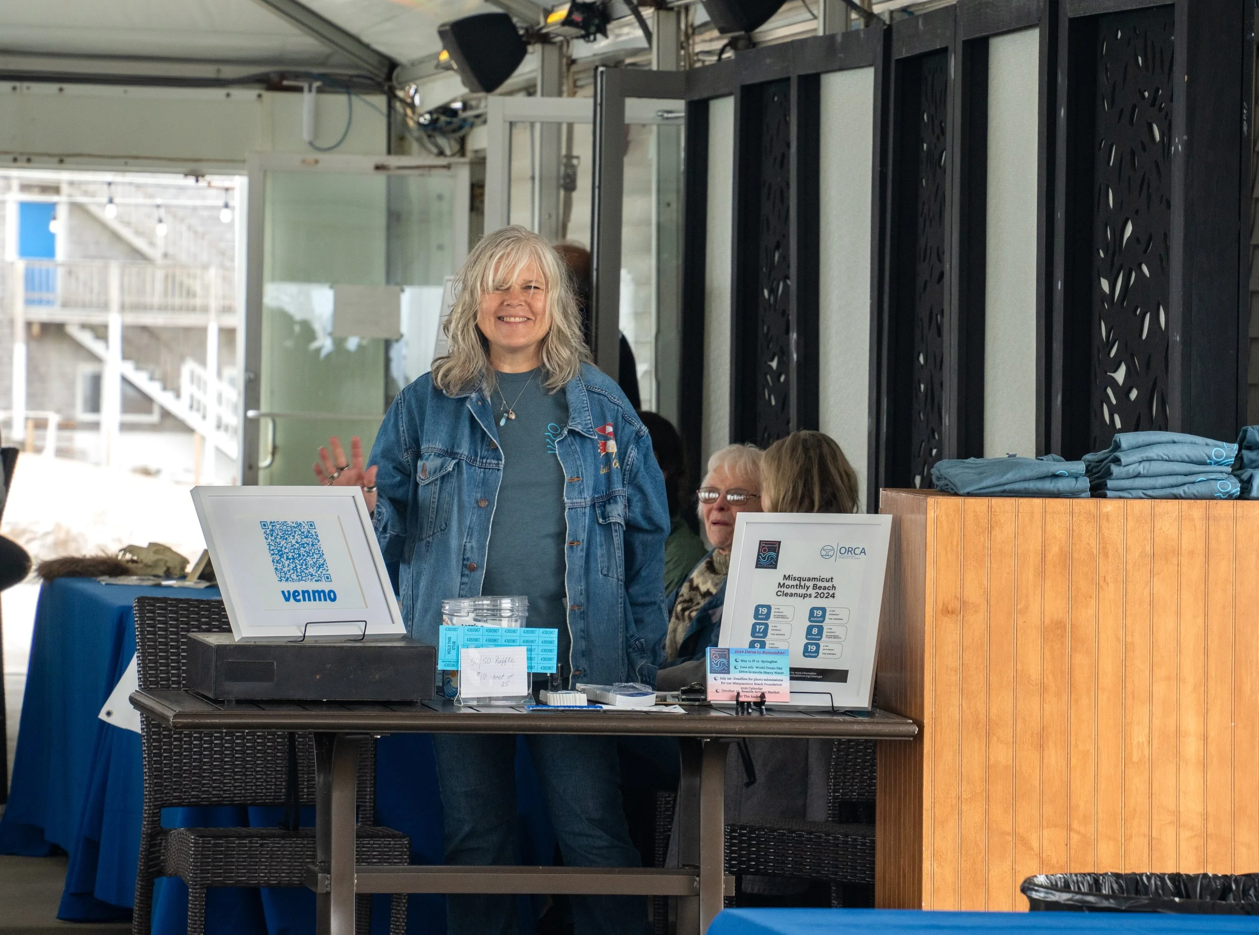 A woman smiling and waving at a booth with raffle tickets and signage for the Misquamicut Artisan Market, in an indoor or semi-outdoor setting with other people behind her.