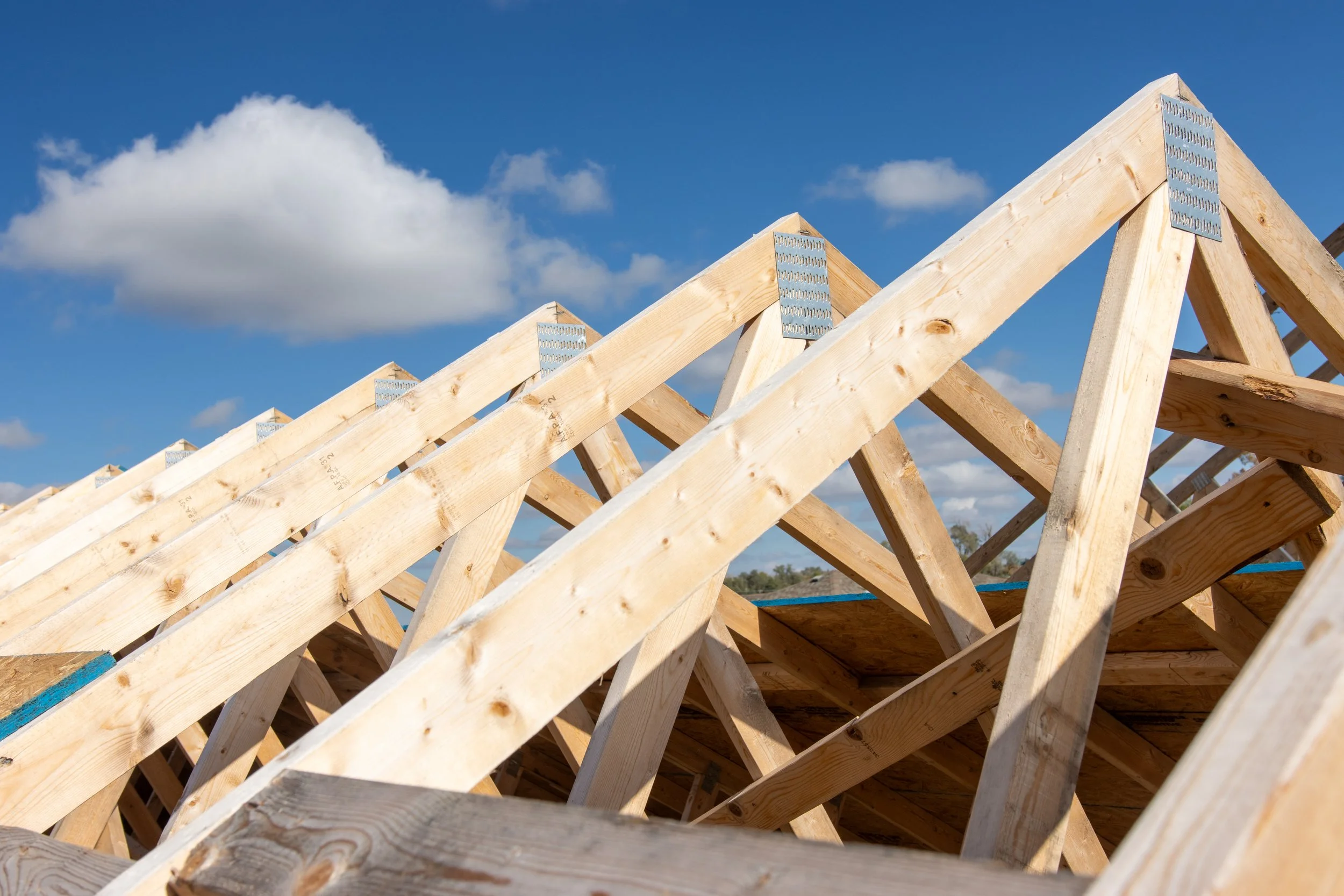 Wooden roof trusses under construction against a blue sky with scattered clouds.