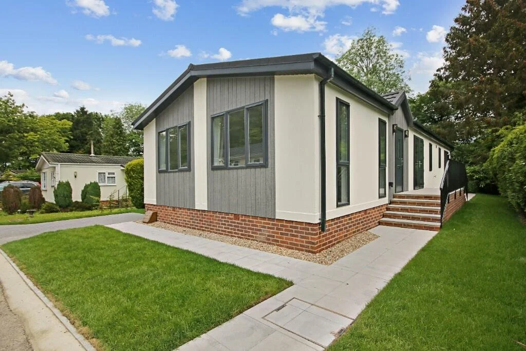 Single-story house with white and gray exterior, brick foundation, black trim, and steps leading to the front door, surrounded by green lawn and trees under a partly cloudy sky.
