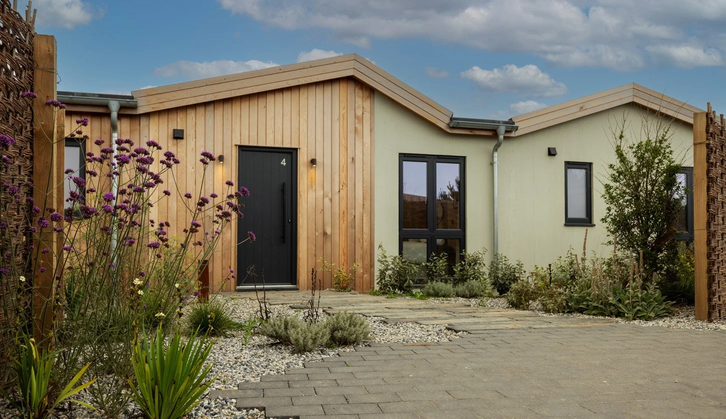 A modern suburban house with a combination of wooden and painted exterior walls, black front door, and large windows, surrounded by a landscaped yard with flowering plants, shrubs, and a stone pathway.