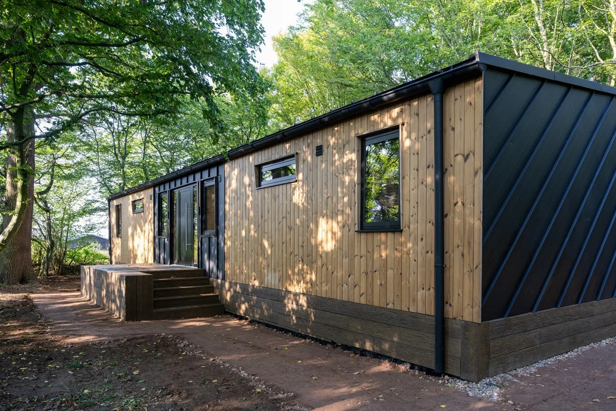 Modern tiny house with a wooden and black exterior, surrounded by trees in a wooded area, with a small set of stairs leading to the entrance.