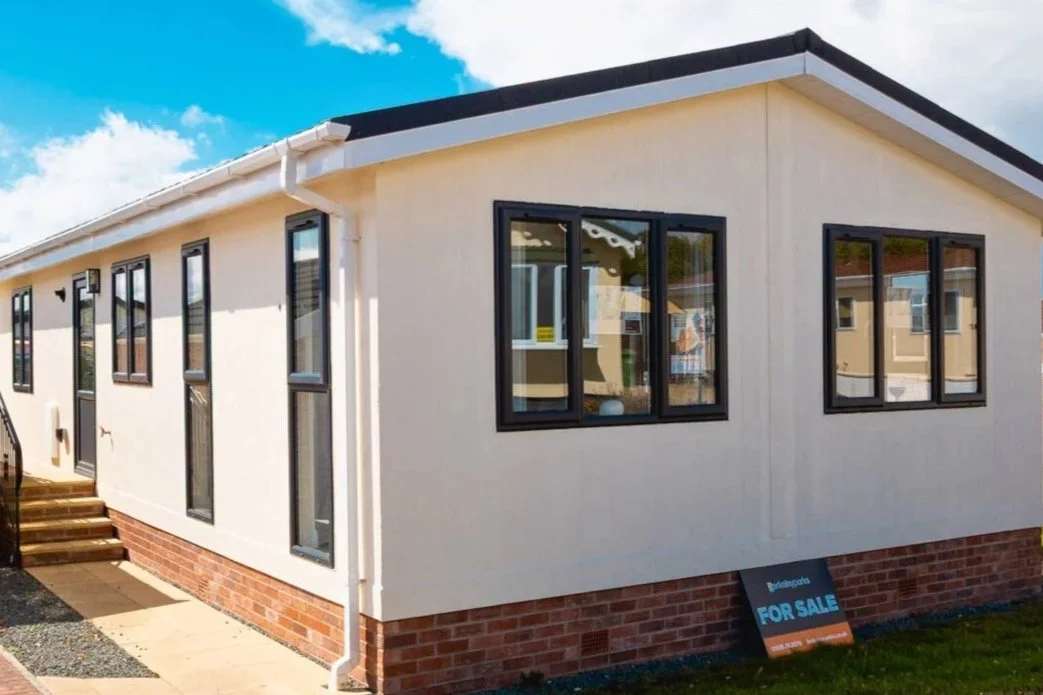 A newly built house with white exterior walls, black window frames, a brick foundation, and a "For Sale" sign outside.
