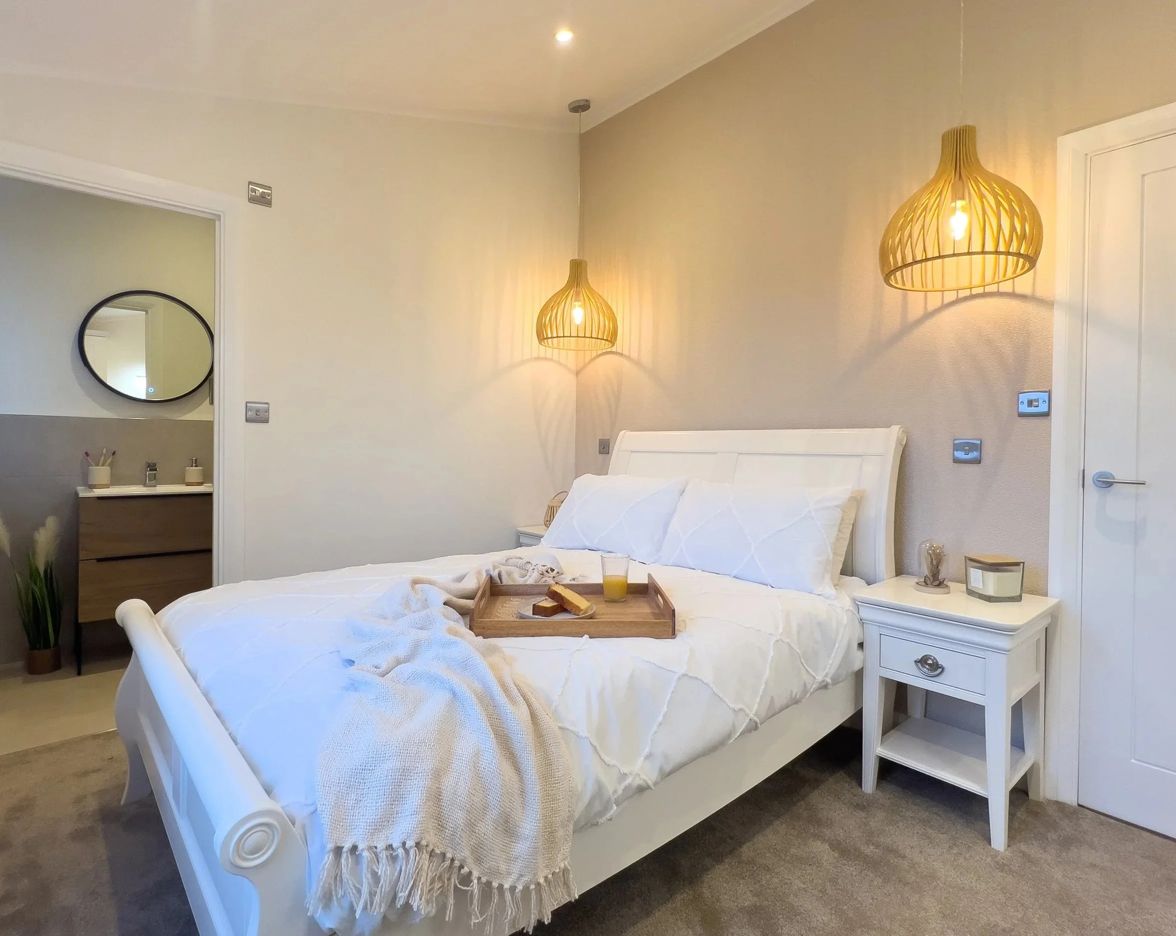 A cozy bedroom with a white bed, two hanging wicker pendant lights, a nightstand with decor, and a tray with breakfast on the bed.