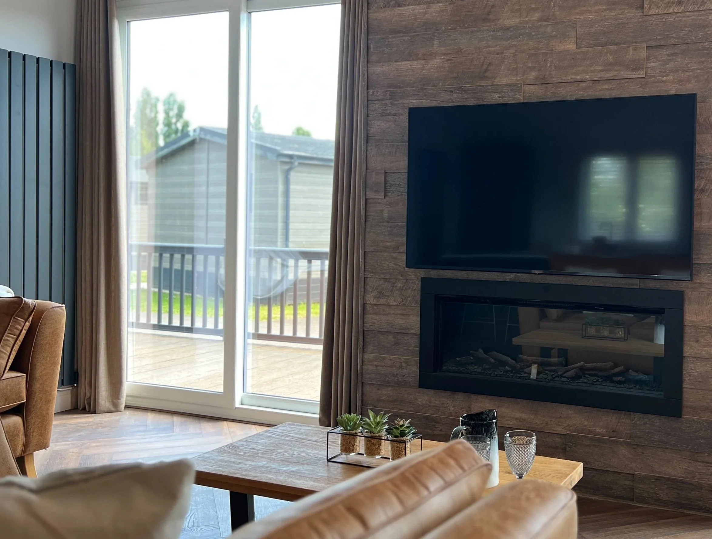 Living room with a large flat-screen TV on a wooden-paneled wall, a fireplace below the TV, a sliding glass door leading to a wooden deck, and a table with small potted succulents and glasses.