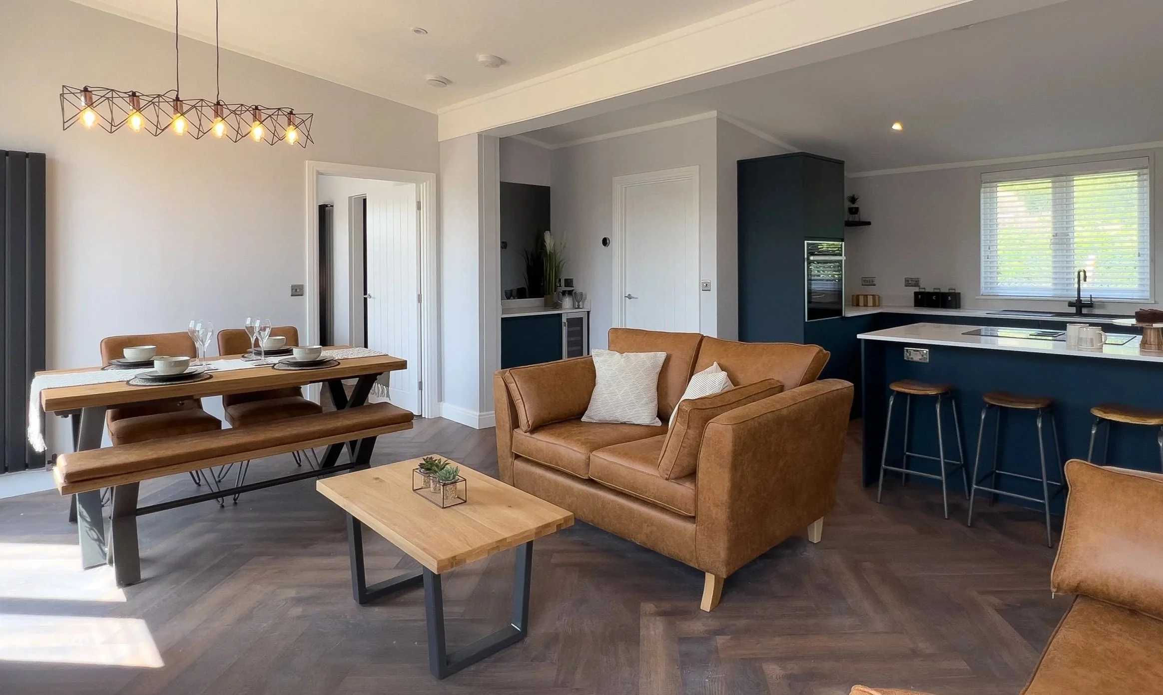 Living room and dining area in a modern open-concept house, with a brown sofa, wooden coffee table, dining table with benches, and a kitchen with bar stools.