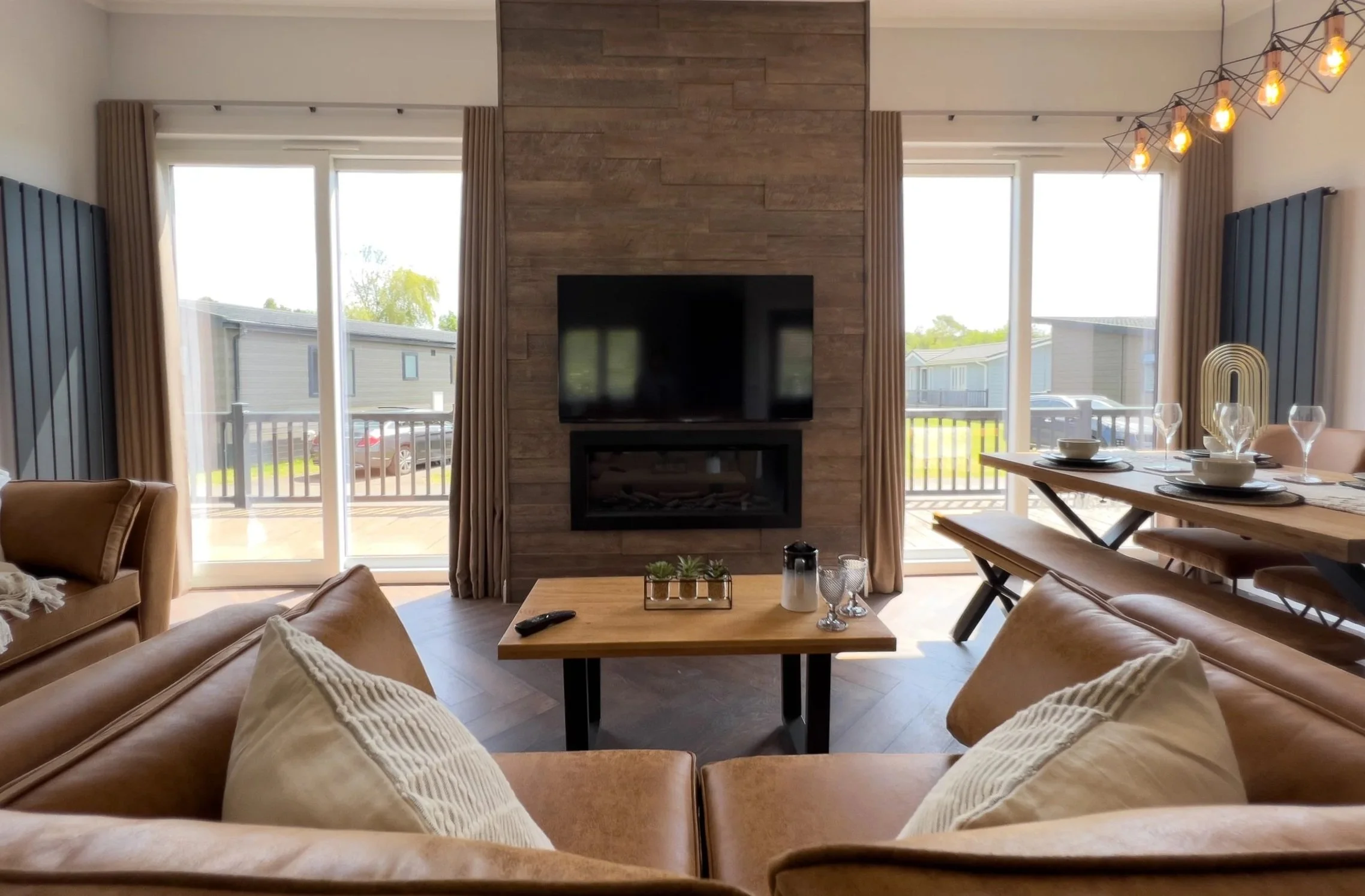 Living room with leather sofas, a central wooden coffee table, a wall-mounted TV over a fireplace, large windows with beige curtains, a dining table with place settings, and decorative lighting.