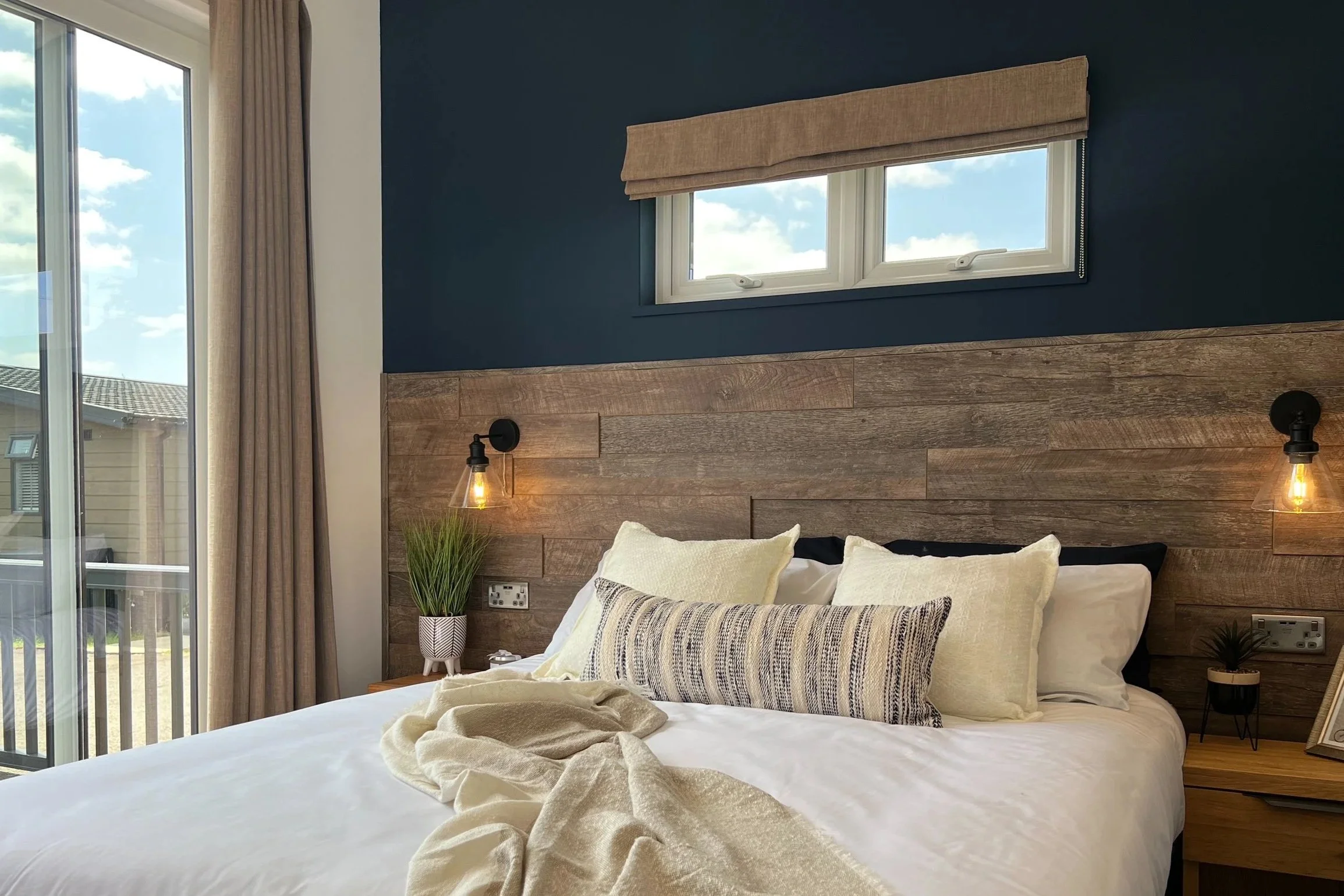 A bedroom with a bed topped with white and patterned pillows, a wooden headboard, two wall-mounted lamps, a small plant on the bedside table, and a window with a beige window covering. Part of a sliding glass door and beige curtains are visible on th
