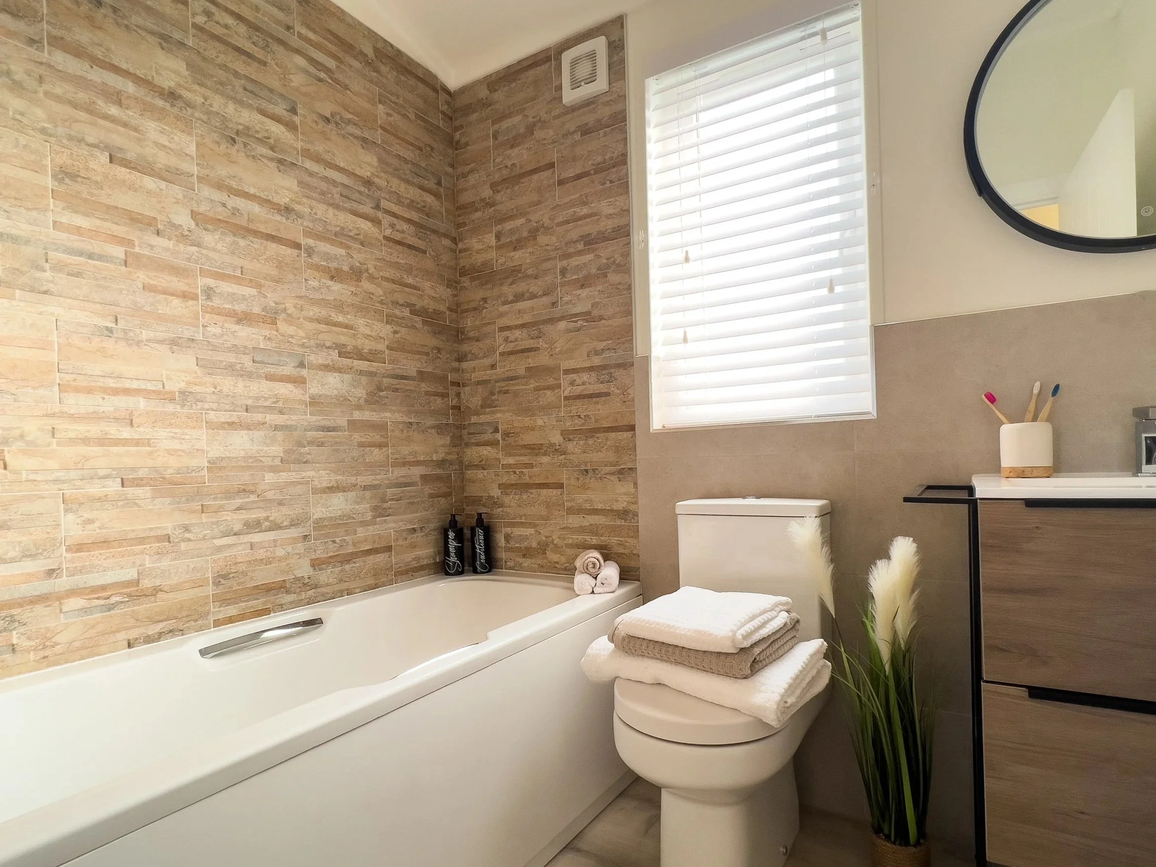 Bathroom with a white bathtub, a toilet with folded towels on top, a wooden cabinet with toothbrushes and a soap dispenser, a window with blinds, a round mirror, and a potted plant with tall, white grass-like leaves.