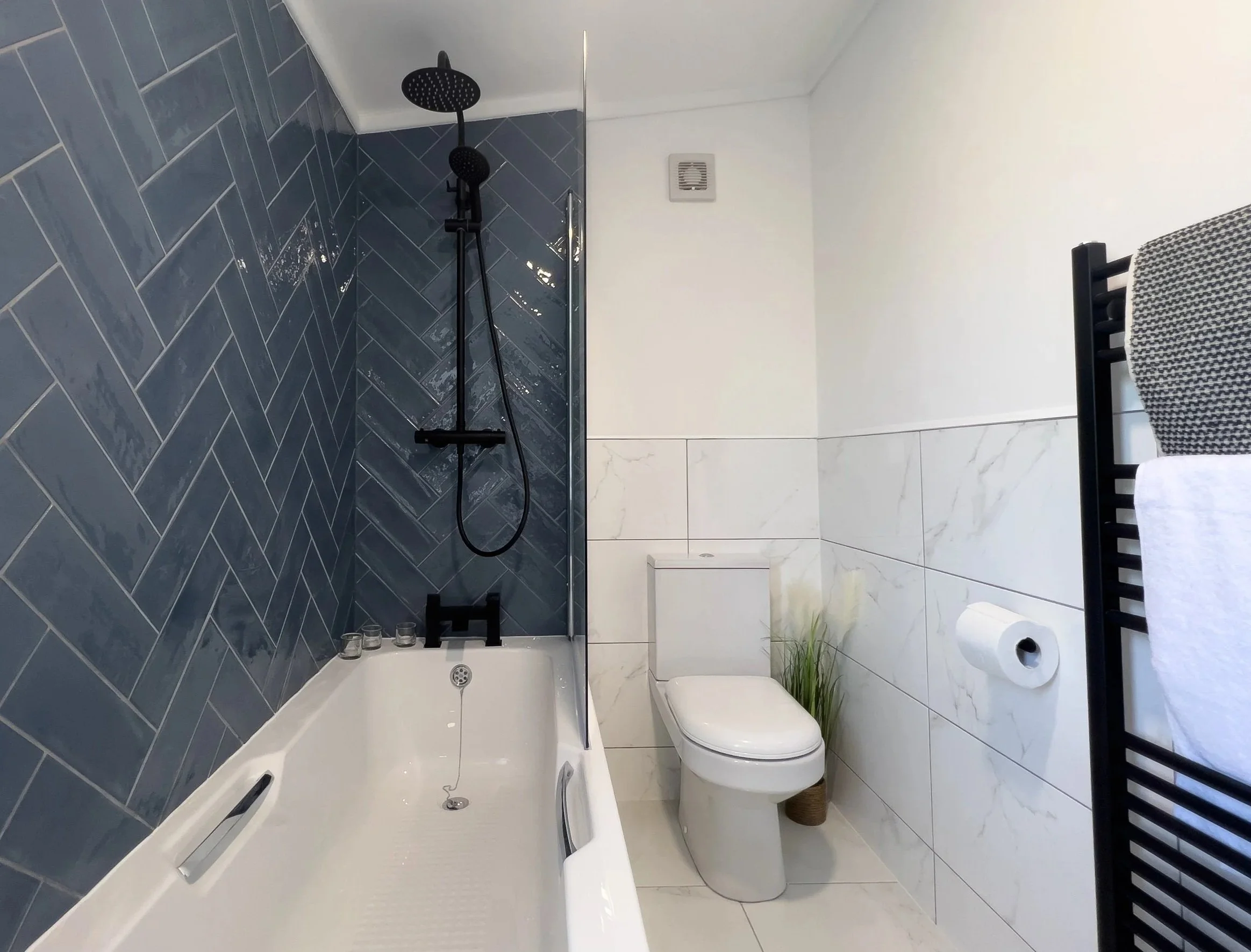 Modern bathroom with a bathtub, black shower fixtures, white toilet with a green plant beside it, and a black towel rack with towels, white and gray herringbone flooring, and white walls with marble tiles.