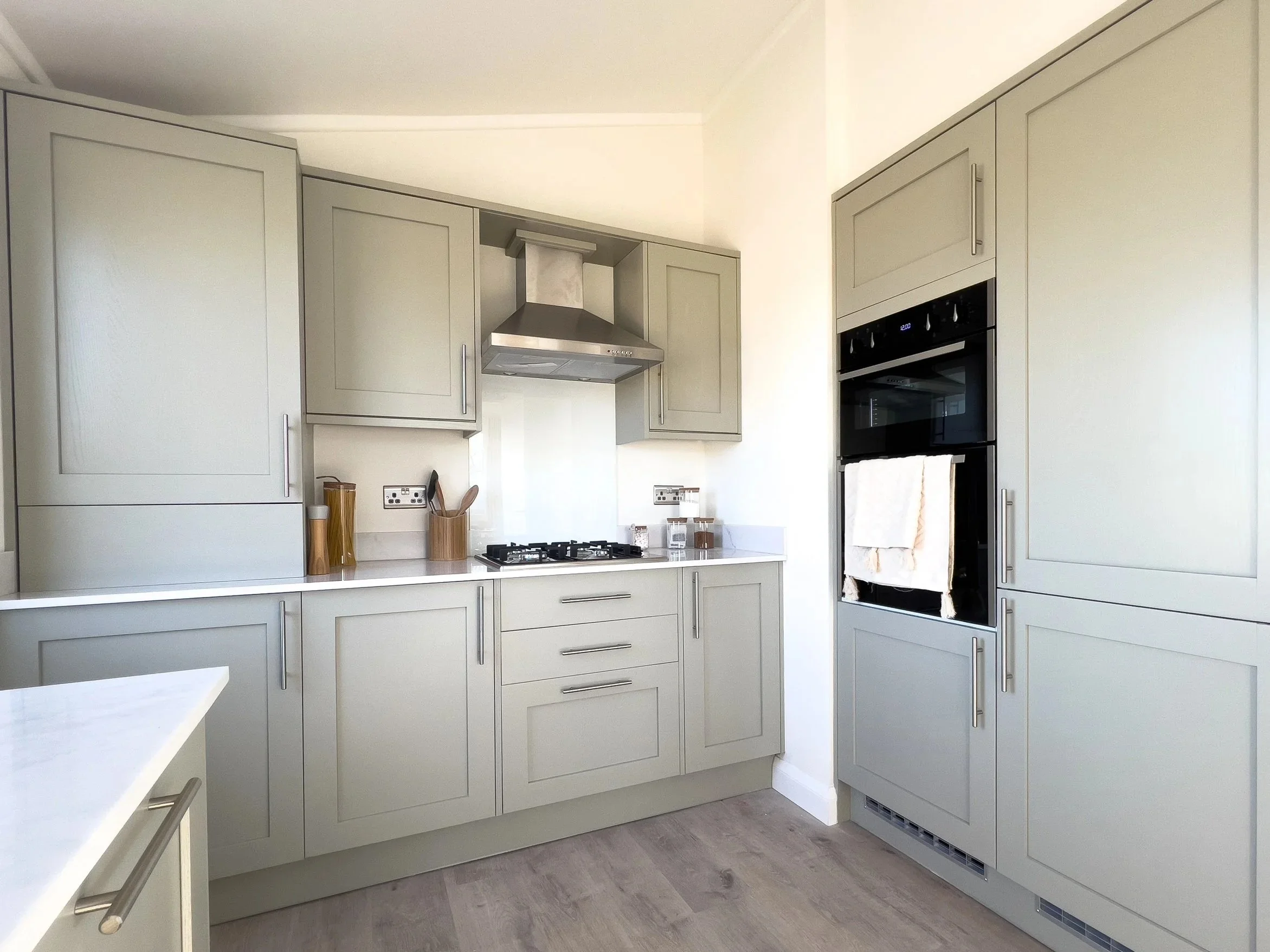 Modern kitchen with light gray cabinets, stainless steel range hood, built-in oven, and a white countertop. A white towel hangs on the oven handle, and there are wooden utensil holders on the counter.