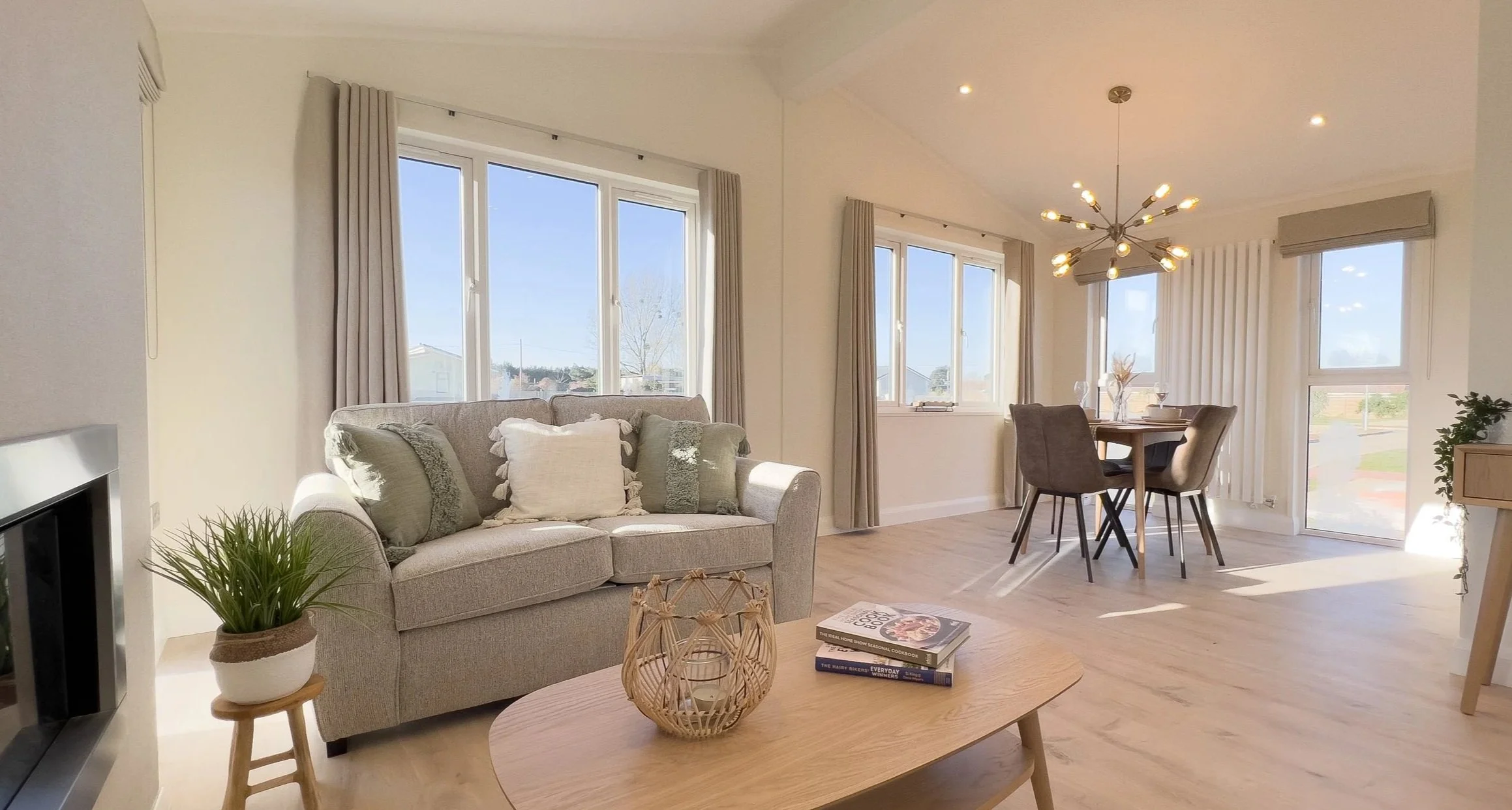 Bright living room and dining area with large windows, beige sofa, wooden coffee table, indoor plant, and modern chandelier.