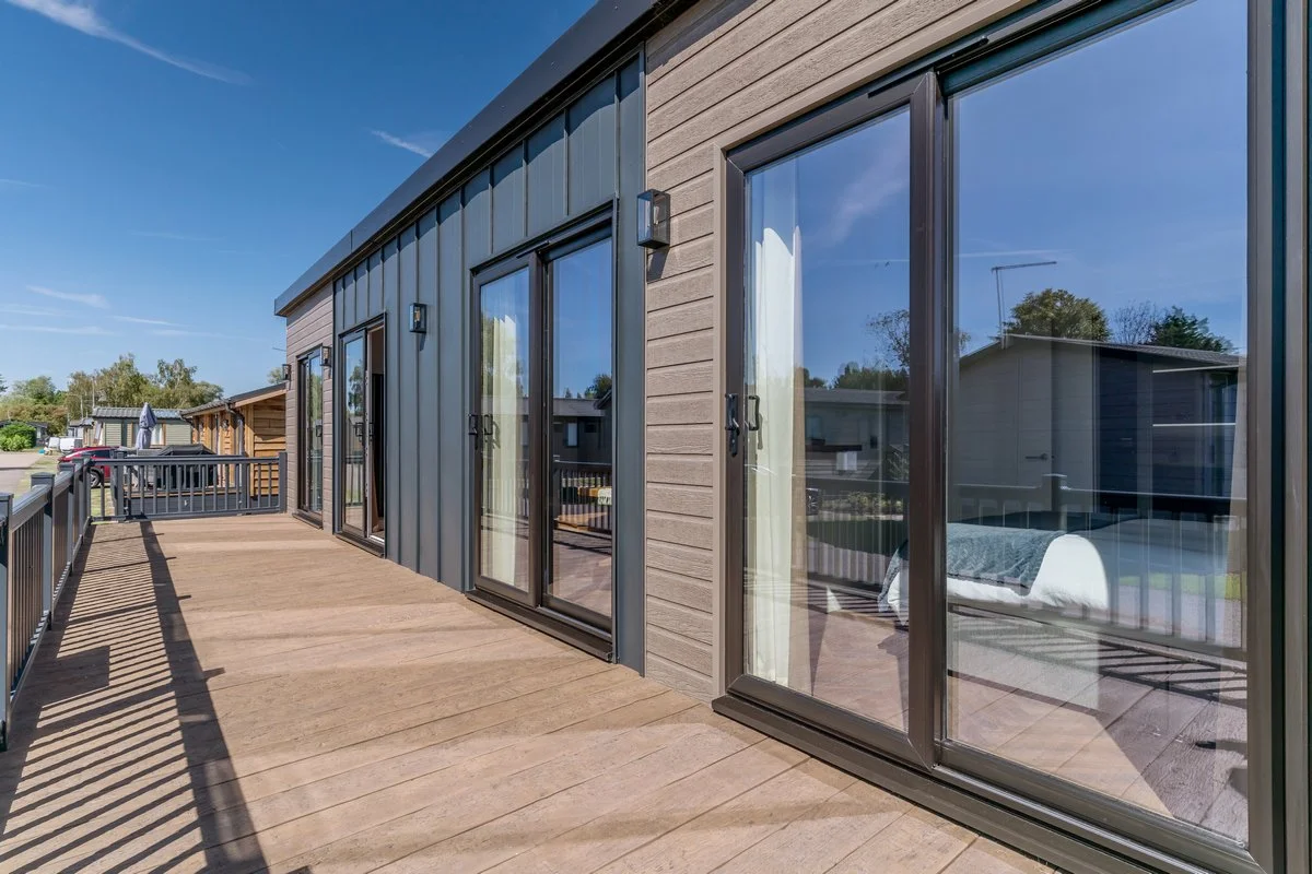 View of a modern house's outdoor deck with wood flooring, metal railing, and glass sliding doors reflecting the sky and neighboring houses.
