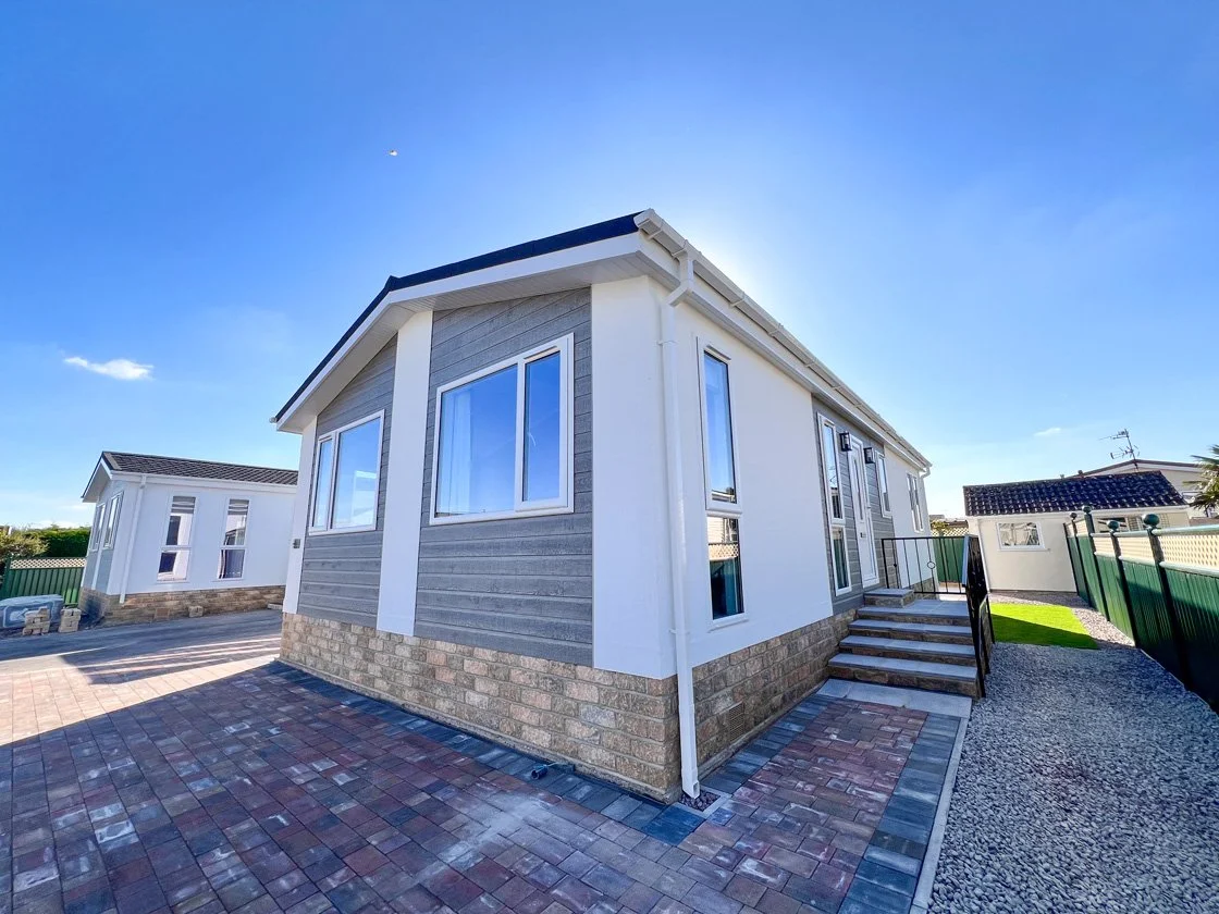 New residential house with gray and white exterior, large windows, small front steps, paved driveway, and fenced yard under clear blue sky.