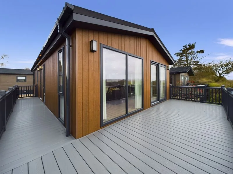 A large wooden house with a spacious gray deck and large glass sliding doors, surrounded by a black railing, with trees and a blue sky in the background.