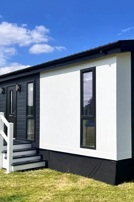 Exterior of a modern tiny house with black and white color scheme, screened porch, black-framed windows, and a sloped roof, set against a blue sky with clouds.