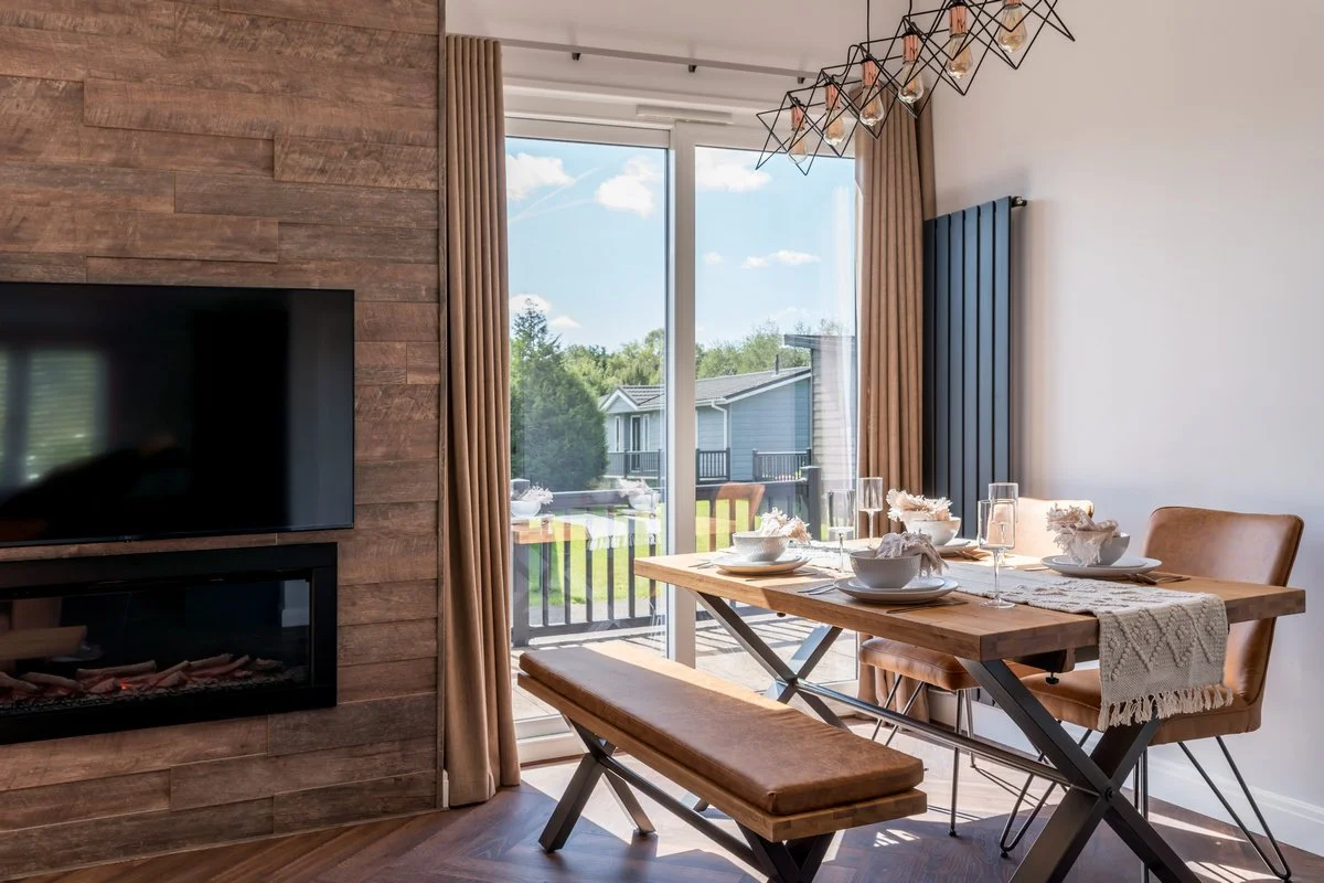 Modern dining area with wooden table, leather chairs, and a bench. Set for a meal with bowls, plates, glasses, and napkins. Large sliding glass door with beige curtains leads to a backyard with houses and trees. A wood-paneled wall with a mounted fla
