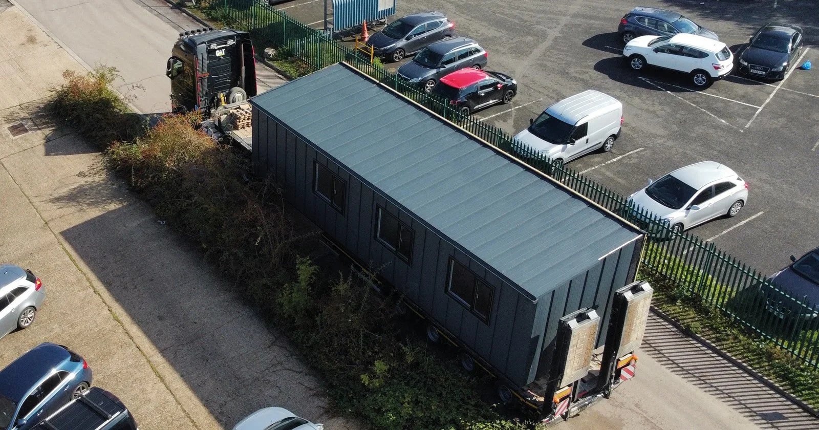 A parking lot with several parked cars and a large mobile home on a trailer being transported. The mobile home is grey with windows, and there are two large units at the back of the trailer. The background shows a fence separating the parking lot from an area with more parked cars and a building.