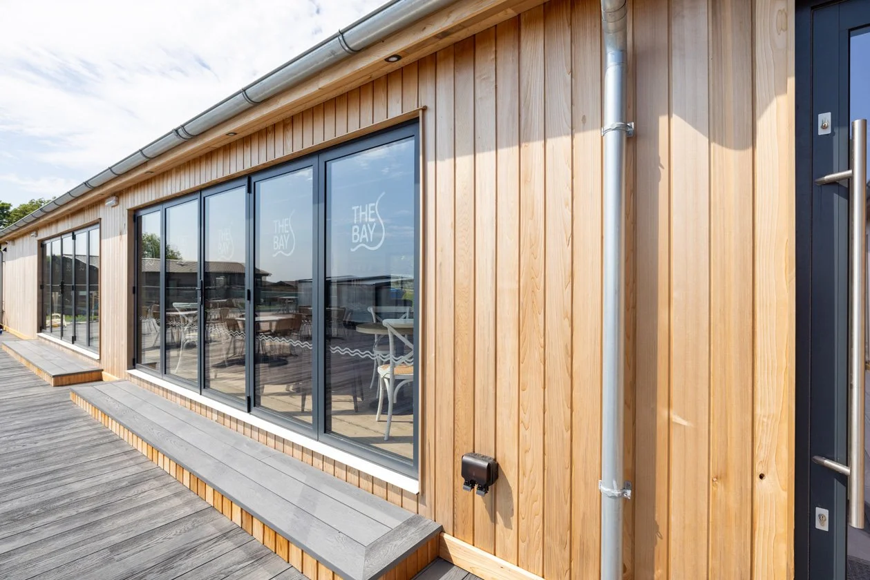 Exterior view of a modern building with wooden paneling, large glass windows, and a wooden deck, possibly a café or restaurant named 'THE BAY' visible through the windows.