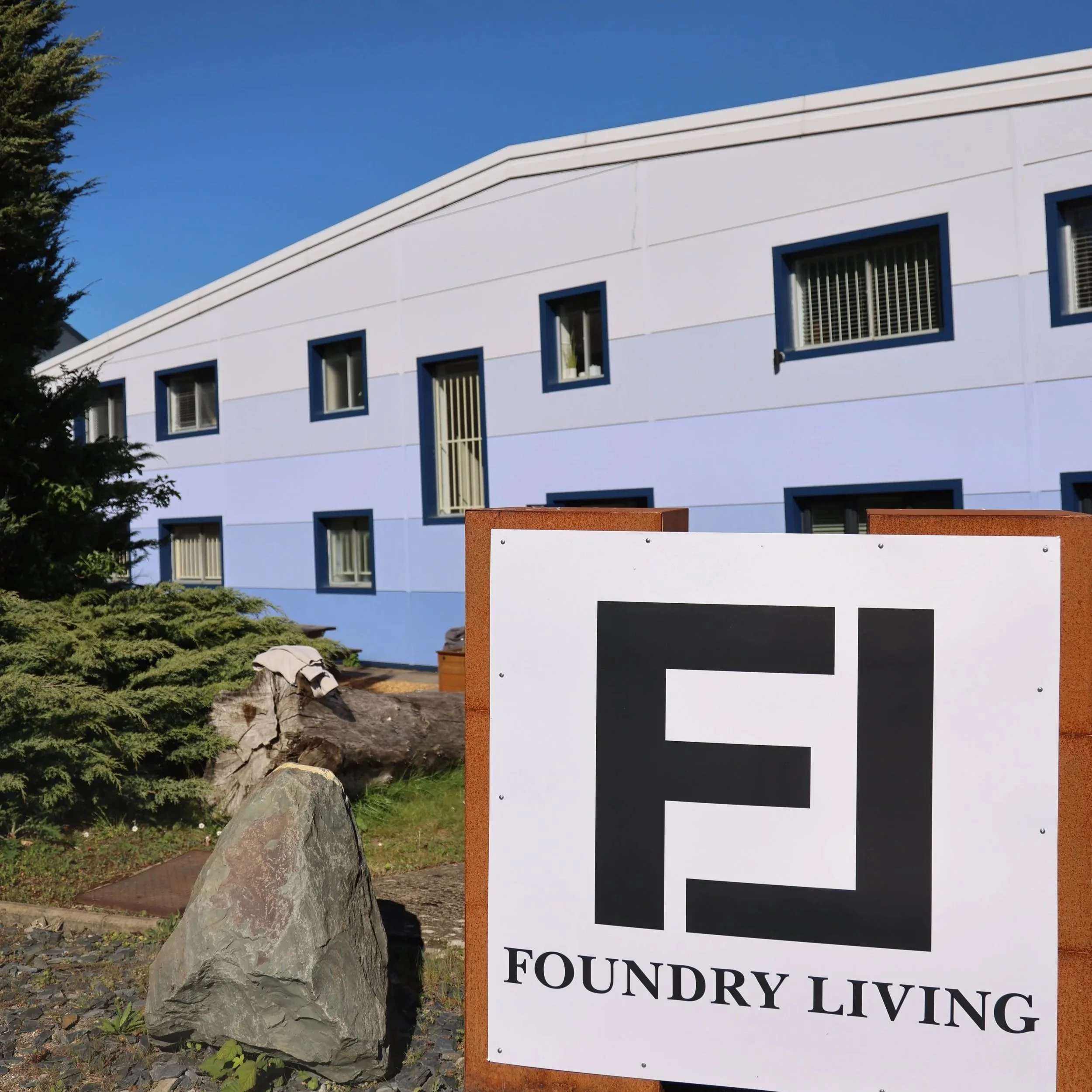 A white building with a blue trim and several small windows, partially obscured by a large tree and rocks. In the foreground, there is a sign with black and white text, reading 'FOUNDATION LIVING,' mounted on a rust-colored frame.