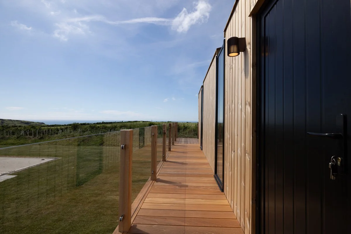 A wooden outdoor balcony with glass railing, attached to a modern building with black and wood exterior, overlooking a grassy landscape and a distant ocean under a partly cloudy sky.