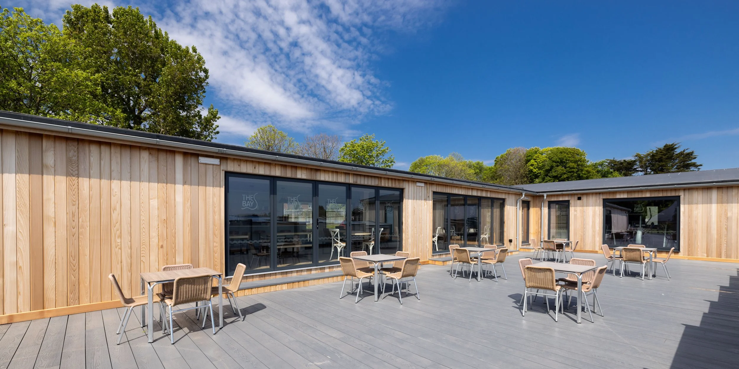 An outdoor patio area with several tables and chairs, next to a modern building with large glass windows and a wooden facade, under a bright blue sky with scattered clouds, surrounded by green trees.