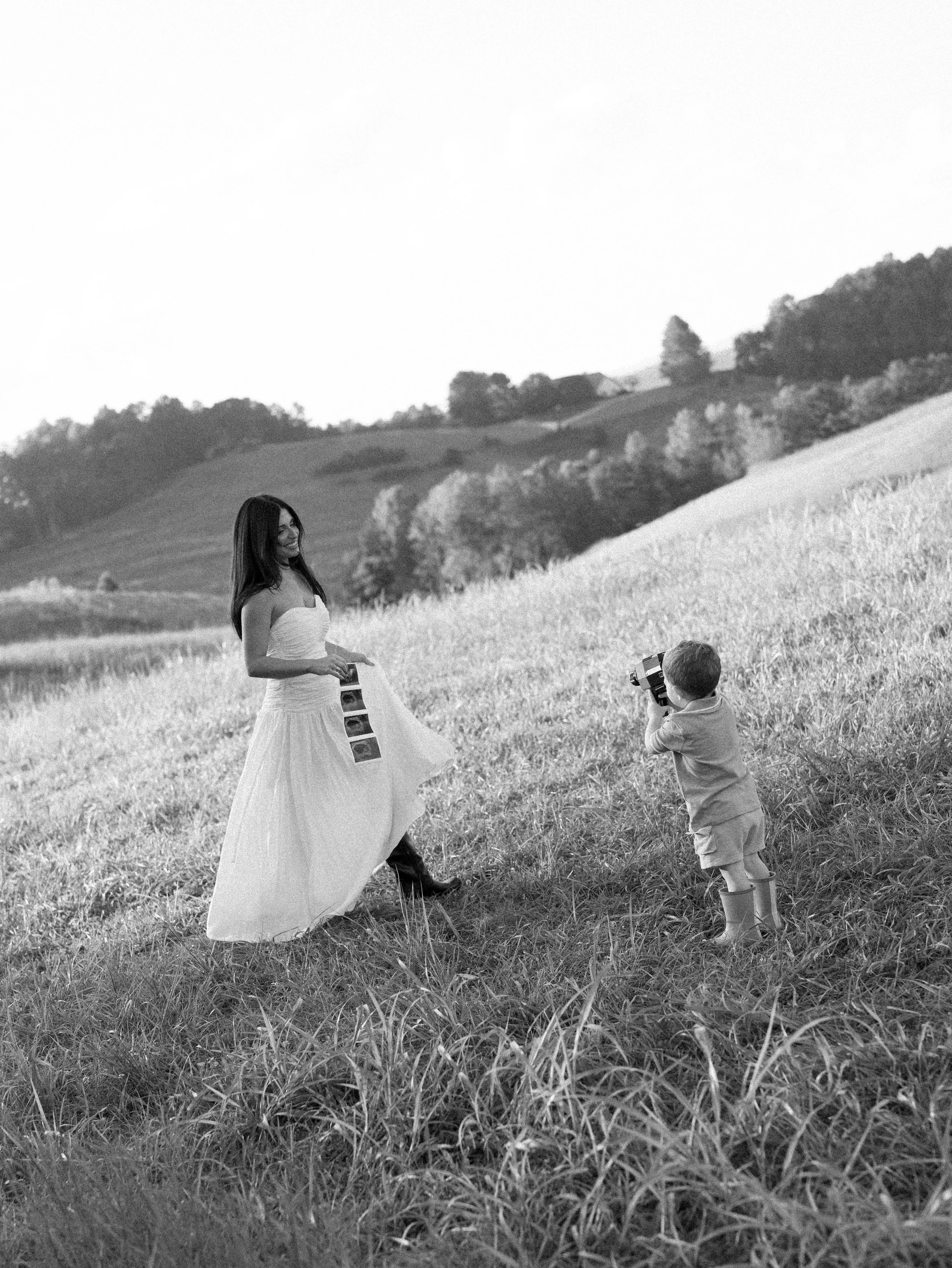 A woman in a strapless dress holding photo strips, smiling while posing in a grassy hill with a boy holding a camera and taking her picture, mountains and trees in the background.