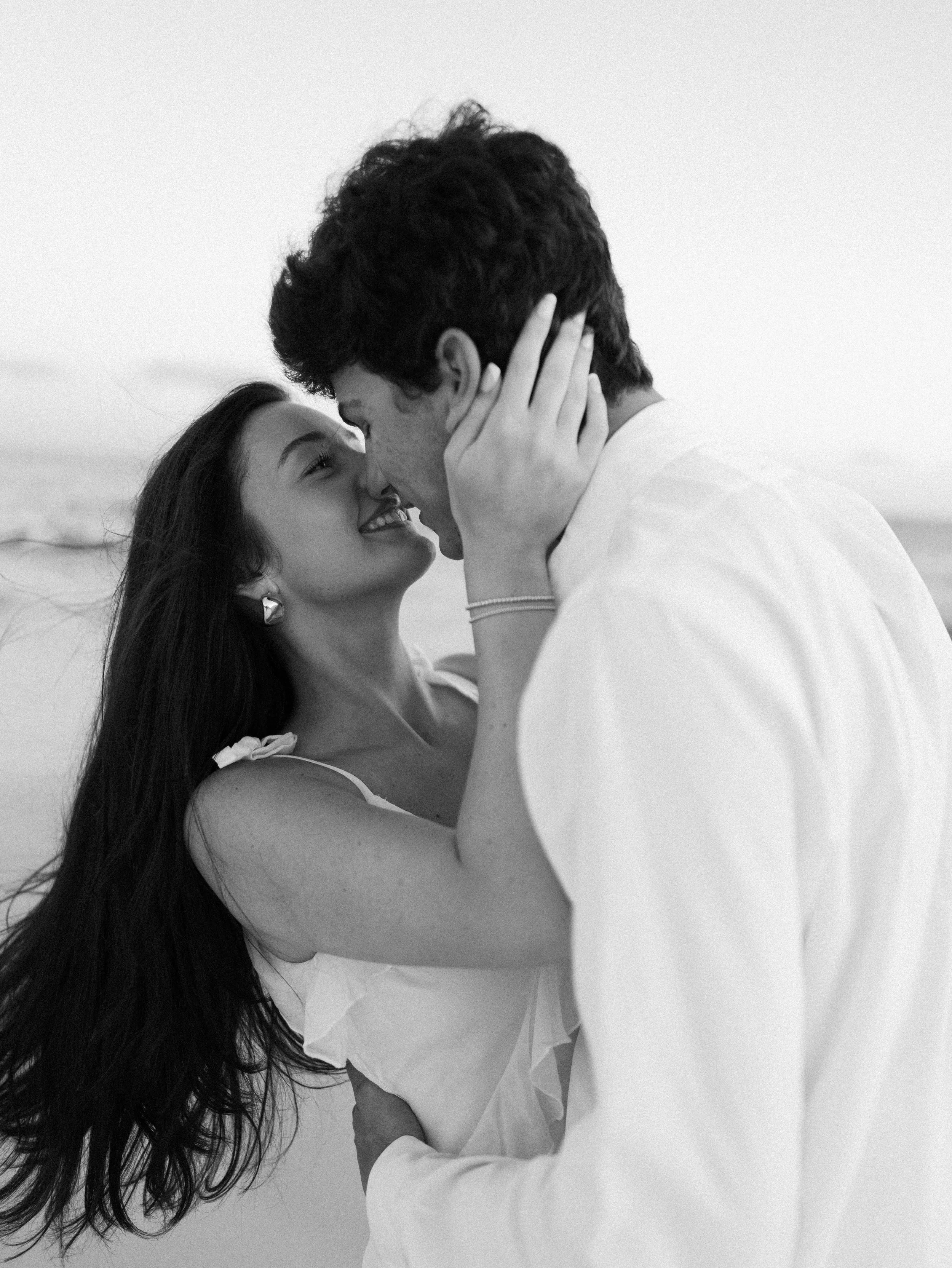 Black and white photo of a young woman and a young man close together, smiling and touching foreheads, in a beach setting with a calm sea in the background.