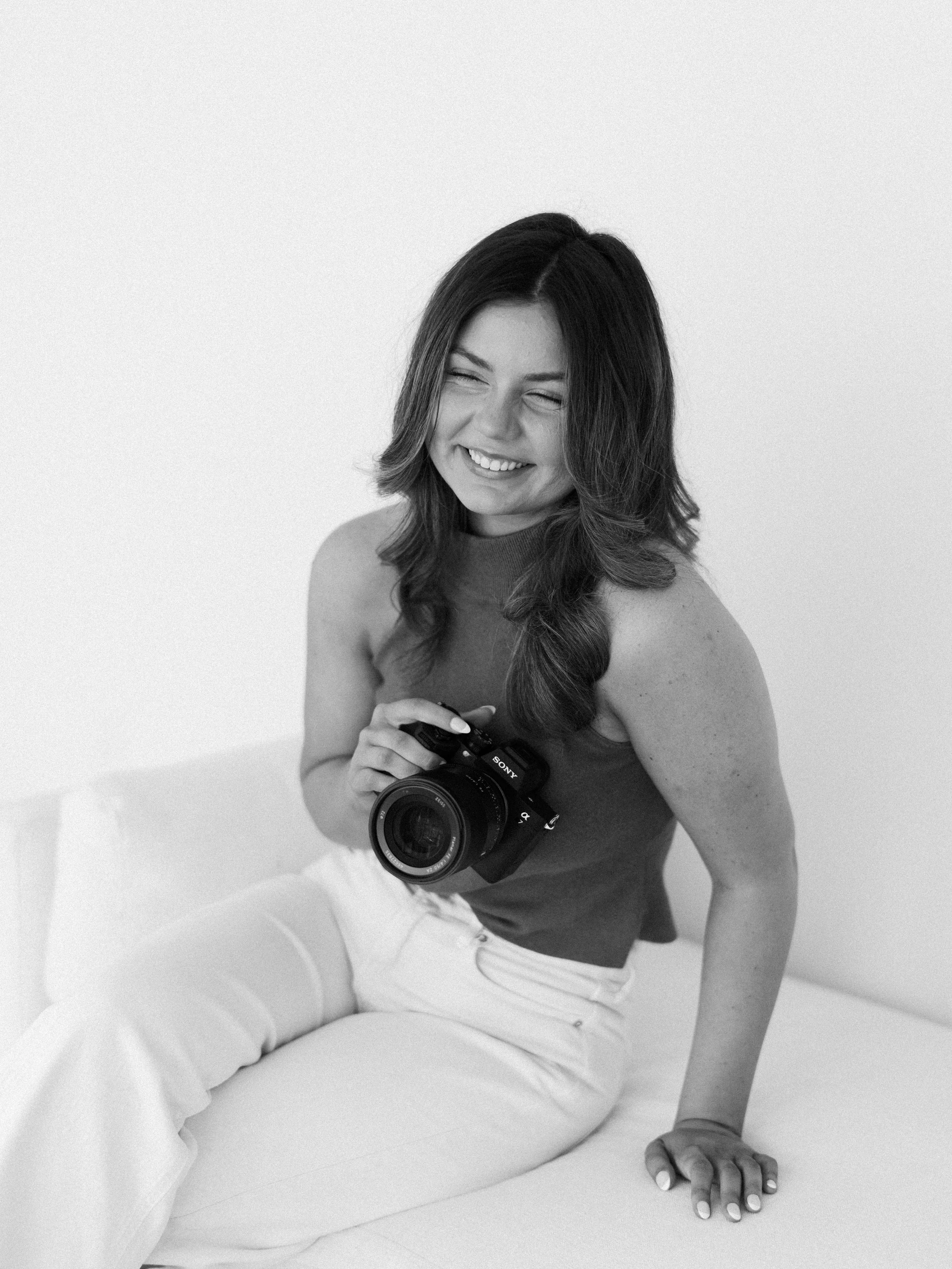 Black and white photo of a smiling woman with long hair, sitting on a white couch, holding a camera, with her right hand on the cushion and her left hand on the couch.