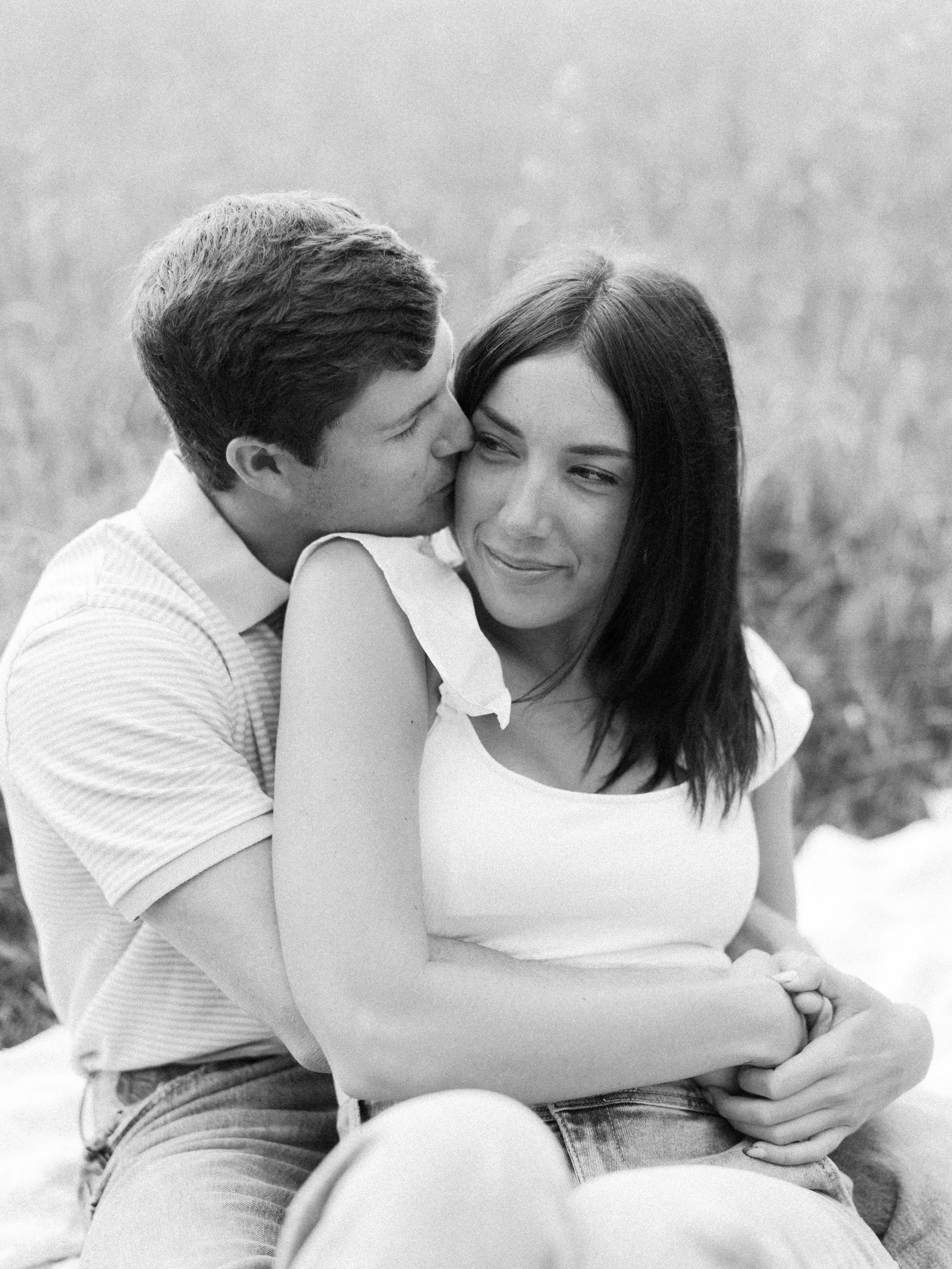 A black and white photo of a man and woman sitting close, with the man kissing the woman's cheek and the woman smiling. They are hugging and outdoors.