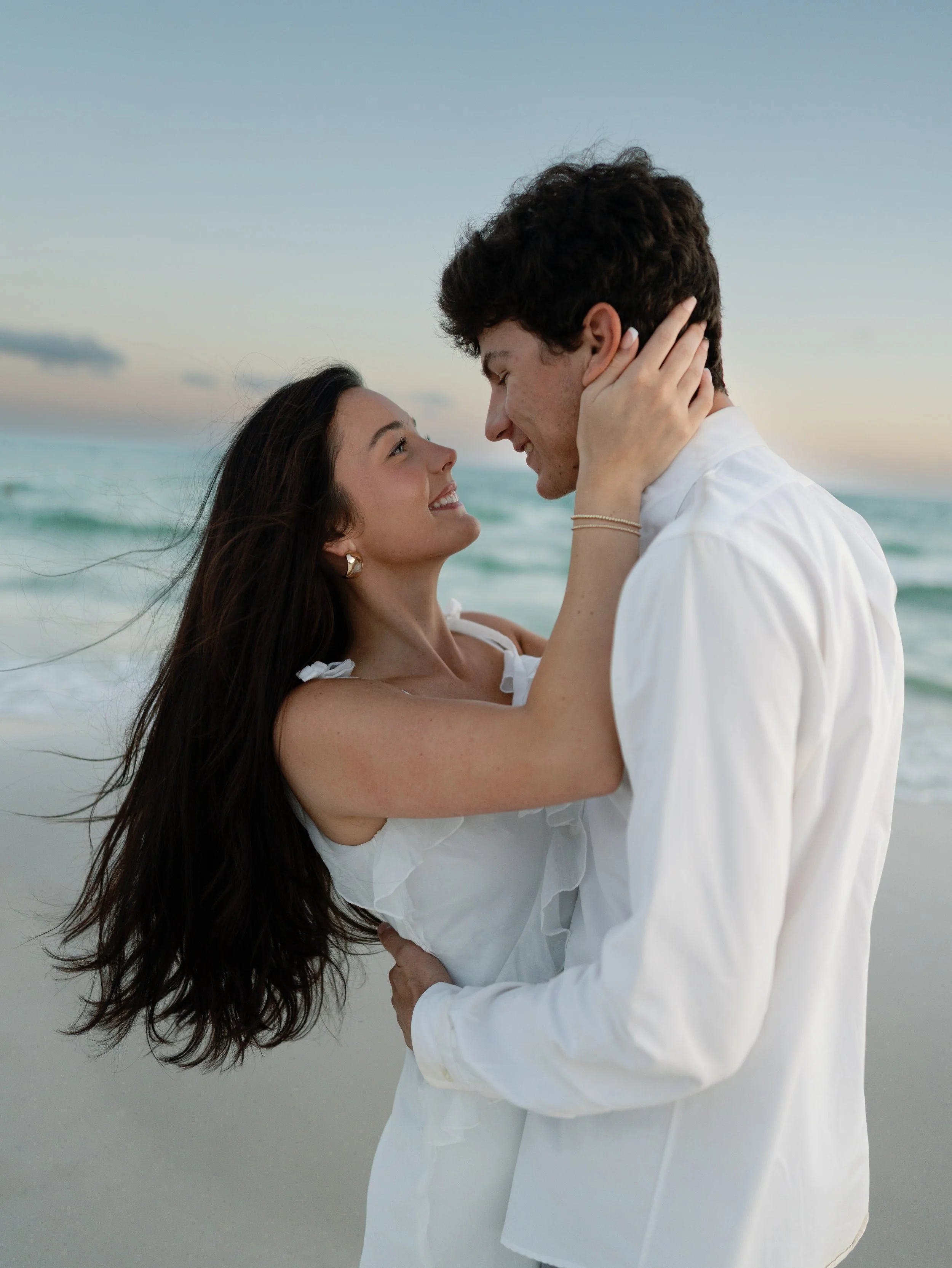 A young couple with light skin at the beach, smiling, with the woman holding the man's face as they look into each other's eyes during sunset.