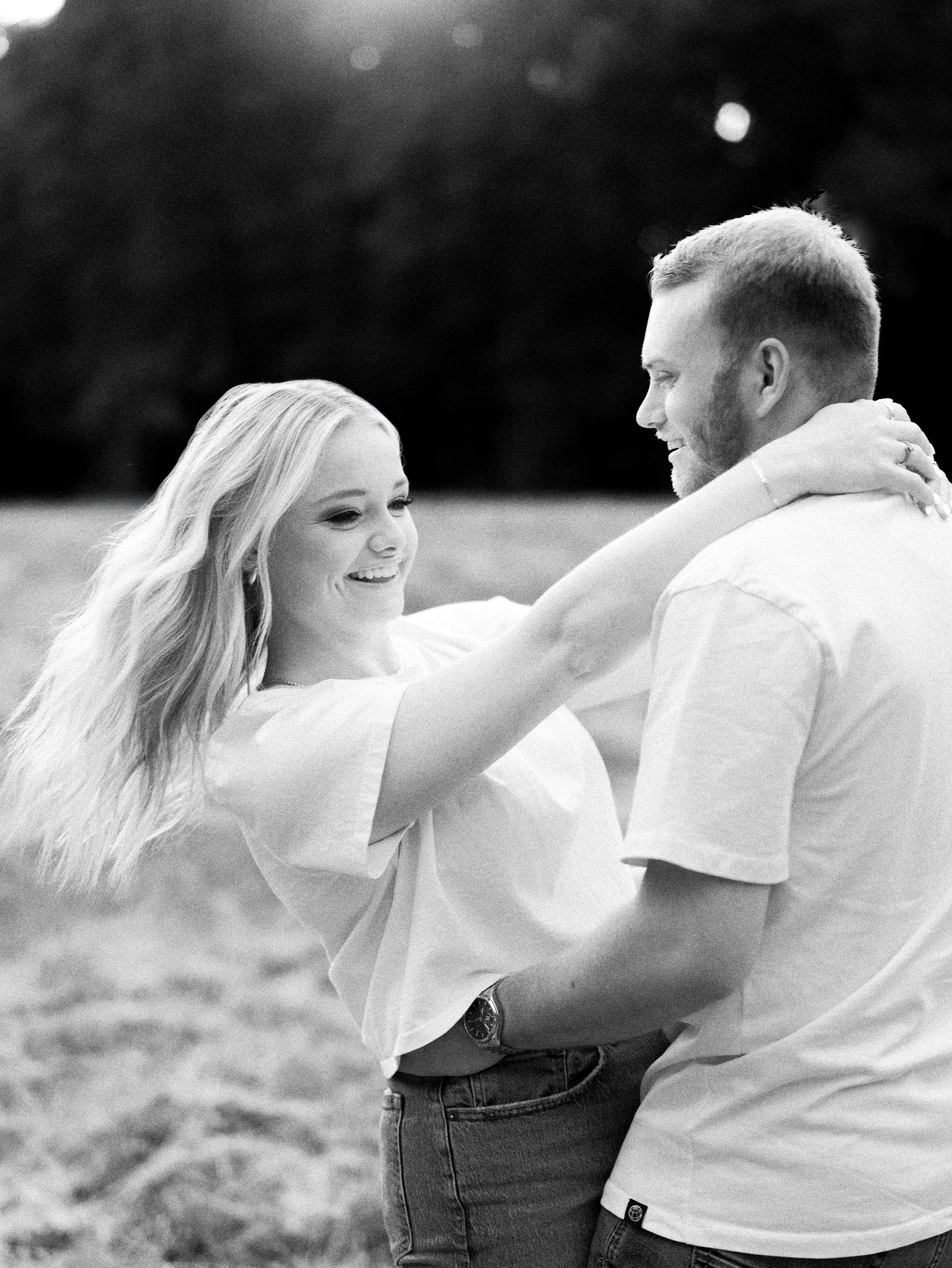 A black-and-white photograph of a couple outdoors, smiling at each other. The woman has long, flowing hair and is wearing a light-colored dress, while the man has short hair, a beard, and is dressed in a light shirt with rolled-up sleeves and jeans, with his arm around her.